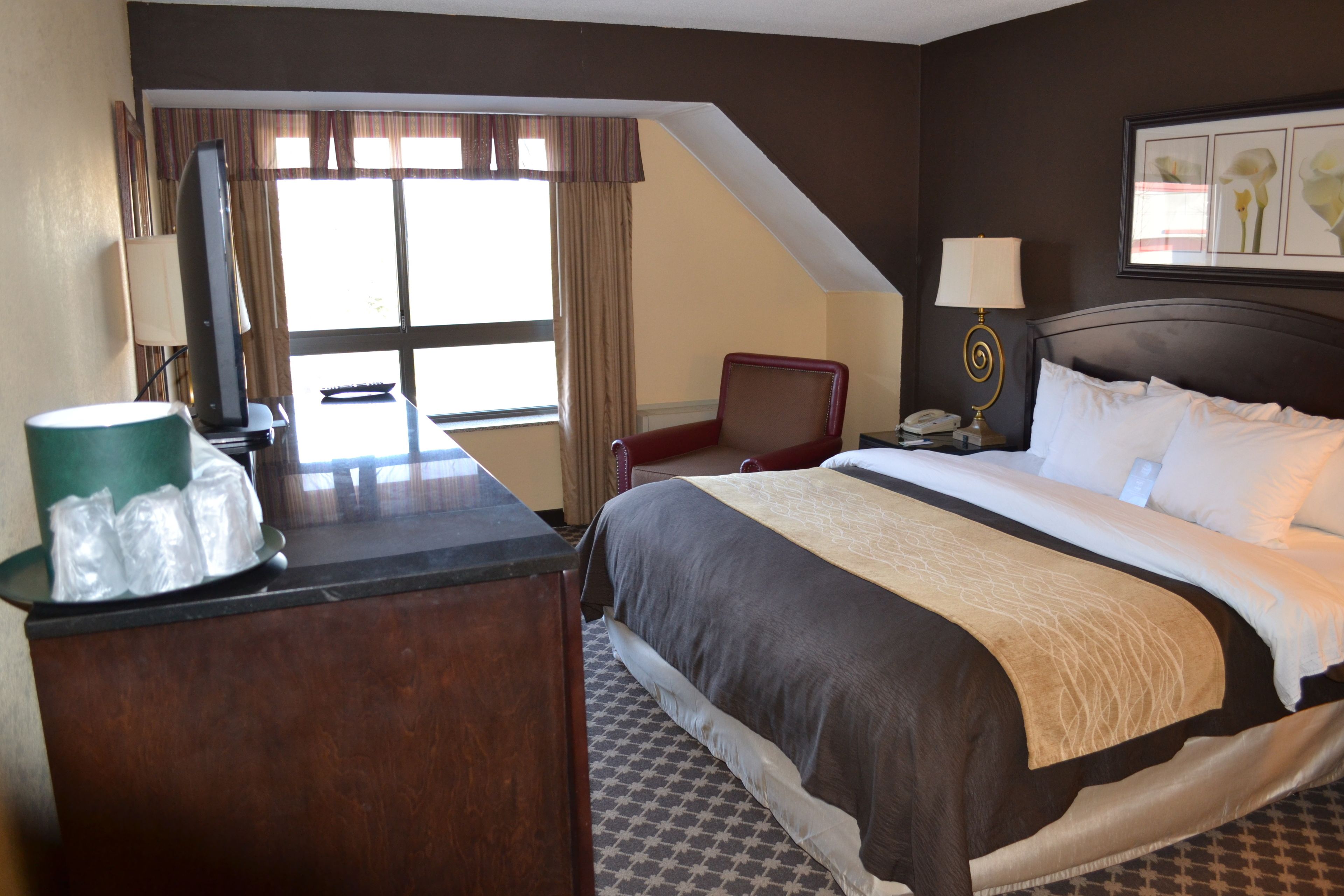 A hotel room with a bed, a desk with a computer, a chair, a window with curtains, and a nightstand with a lamp.