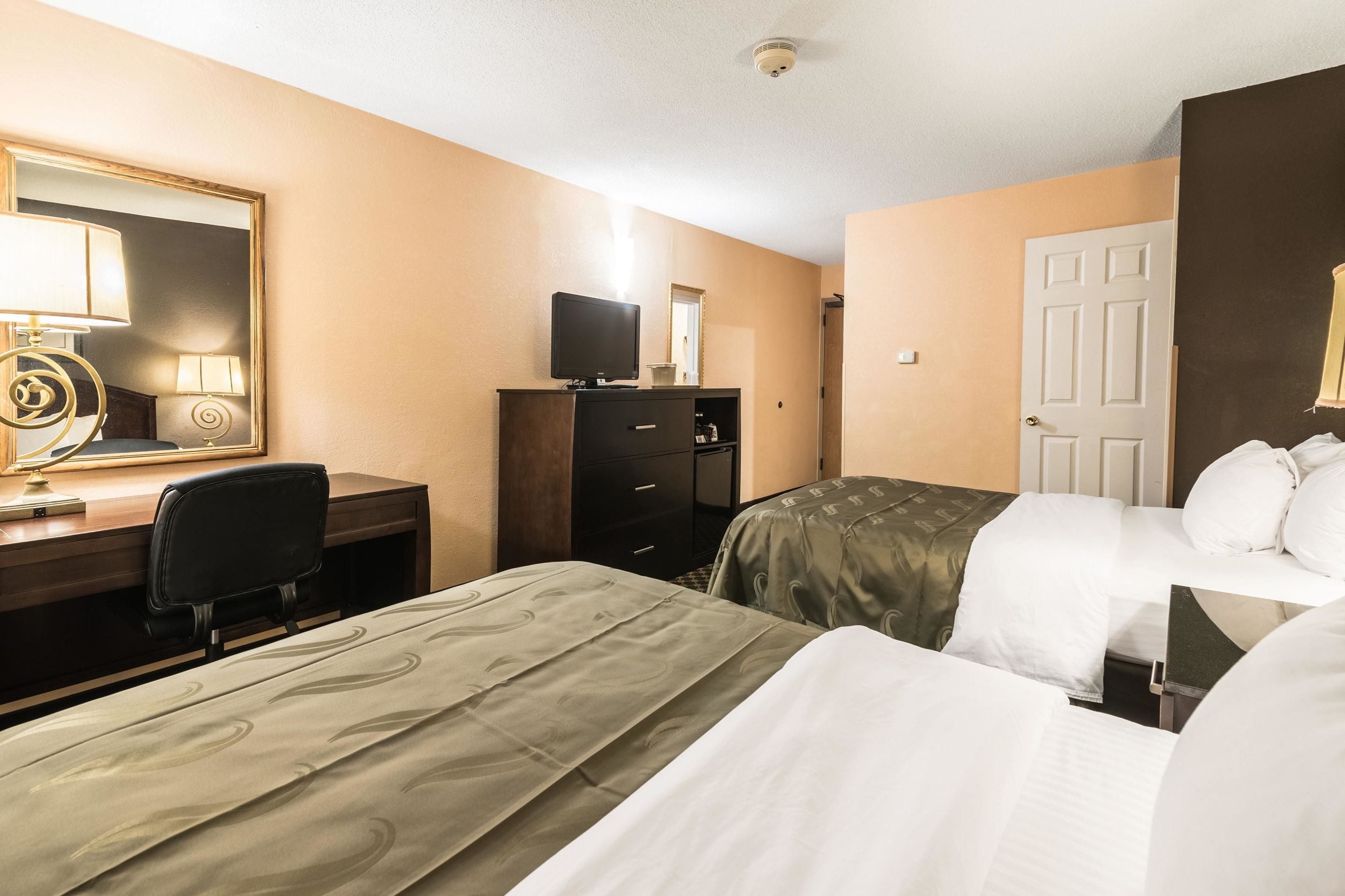 A hotel room with two beds, a desk with a chair, a dresser, a television, and a mirror.