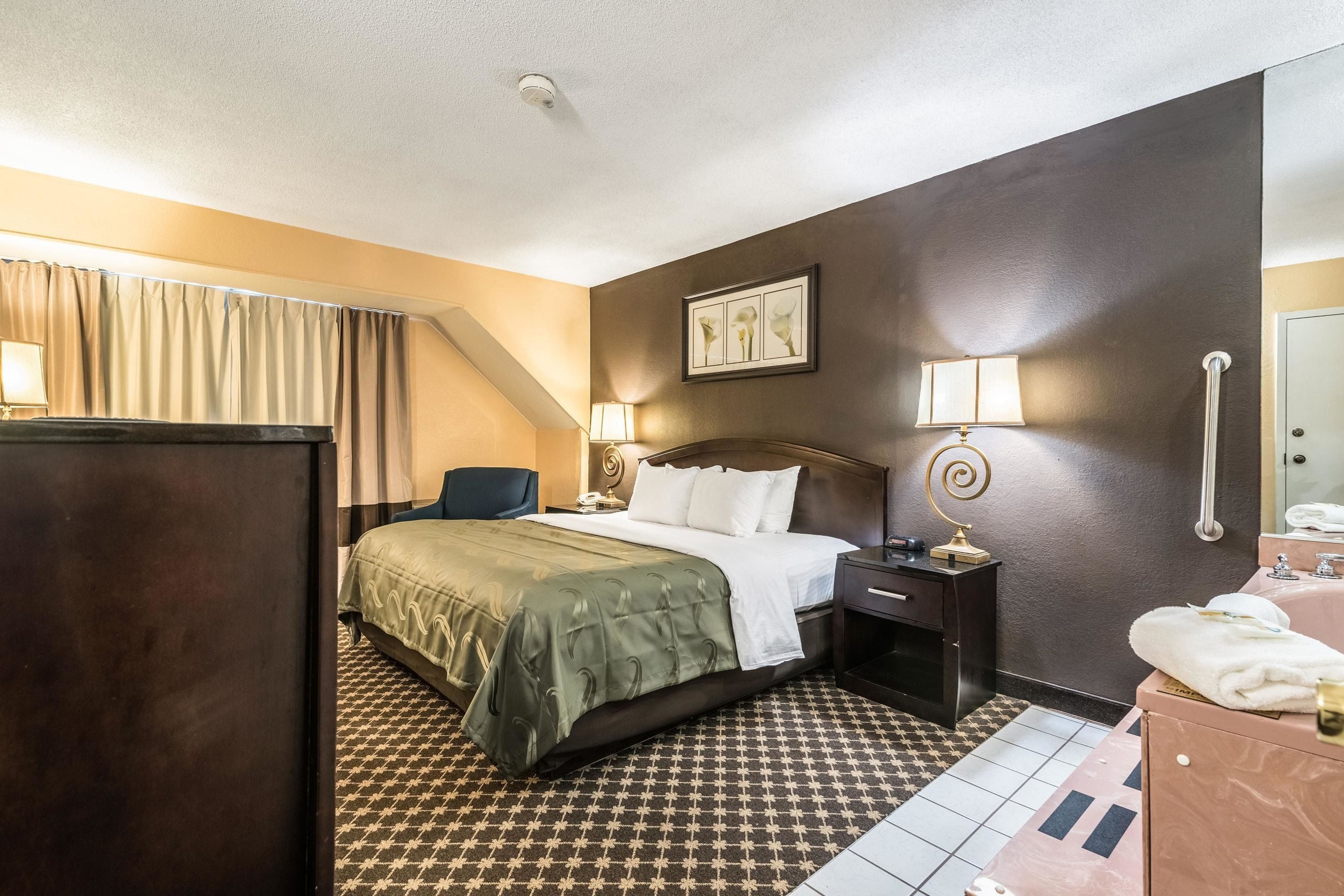 Habitación de hotel con cama, mesita de noche, lámpara y baño con lavamanos y espejo.