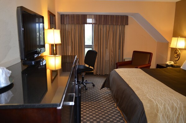 Habitación de hotel con cama, escritorio con televisor, silla, lámpara de escritorio y ventana con cortinas.