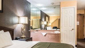 A hotel room with a bed, a nightstand, a lamp, a mirror, a bathroom with a bathtub, and a door.