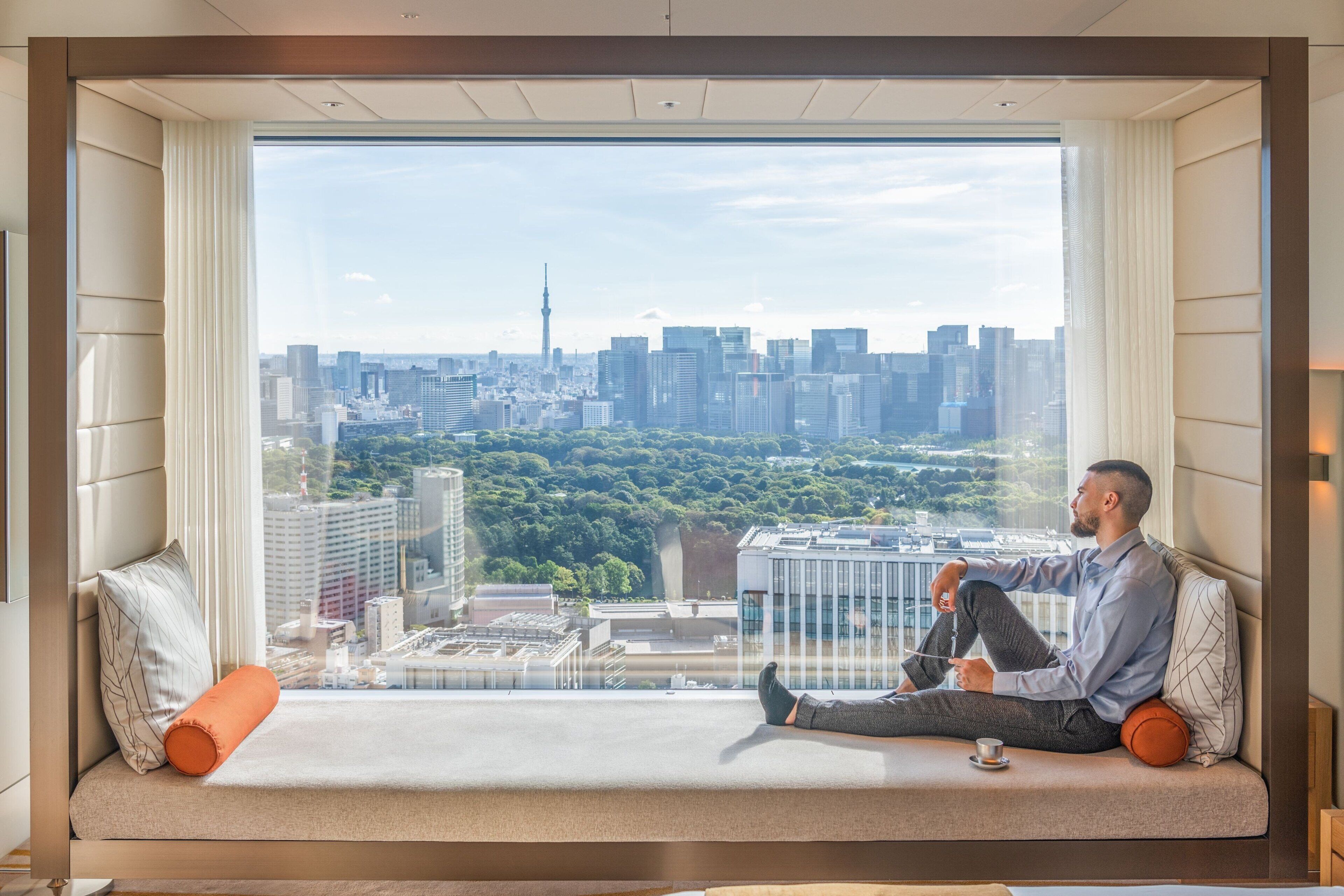 A man sitting on a sofa by a large window with a cityscape view.