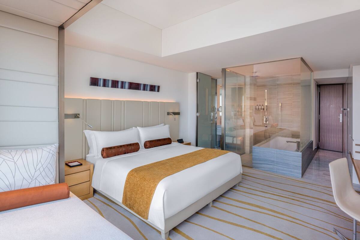 A modern hotel room with a large bed, bedside tables, a glass-enclosed shower, and a small seating area.