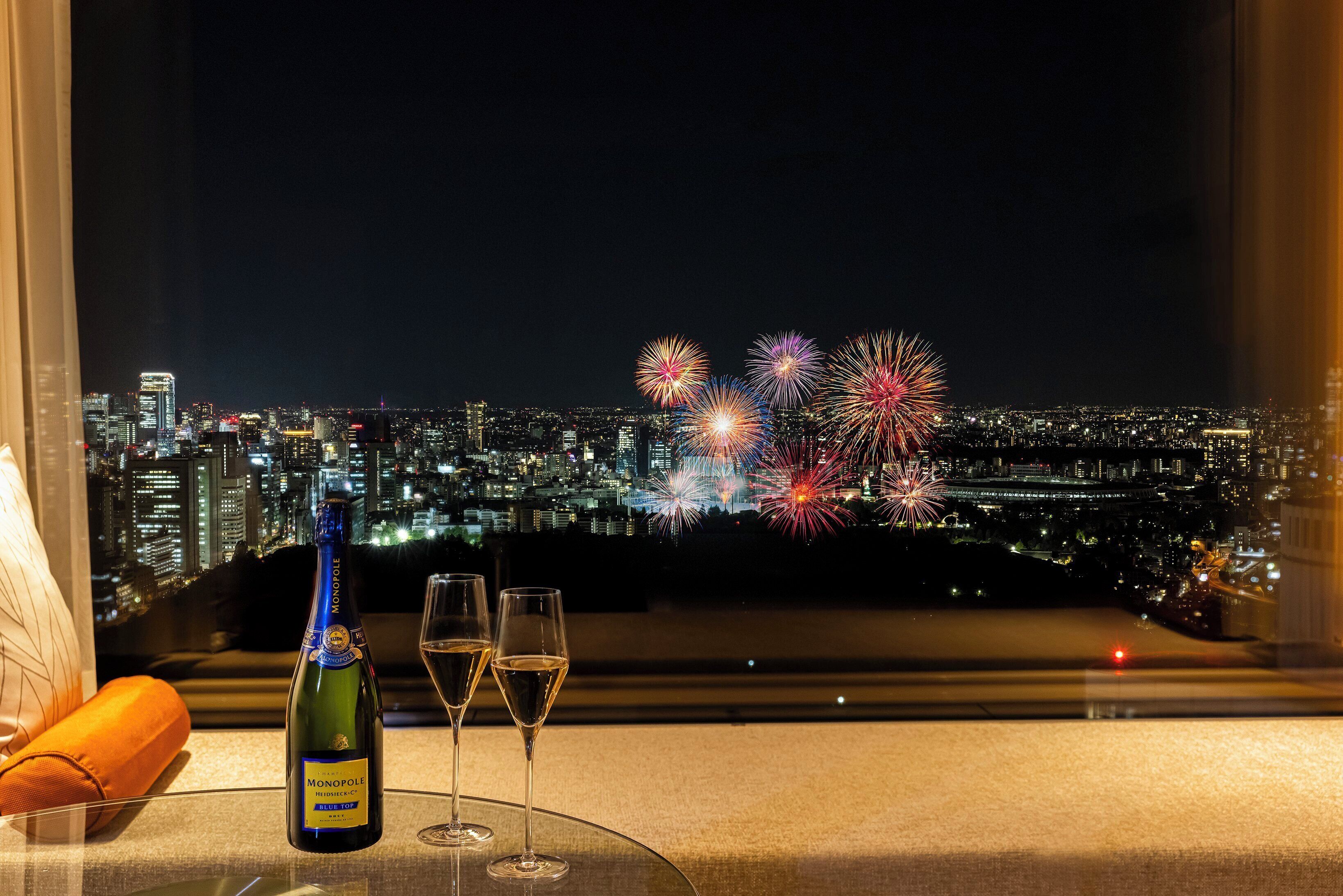 A bottle of champagne and two glasses on a table with a cityscape and fireworks in the background.