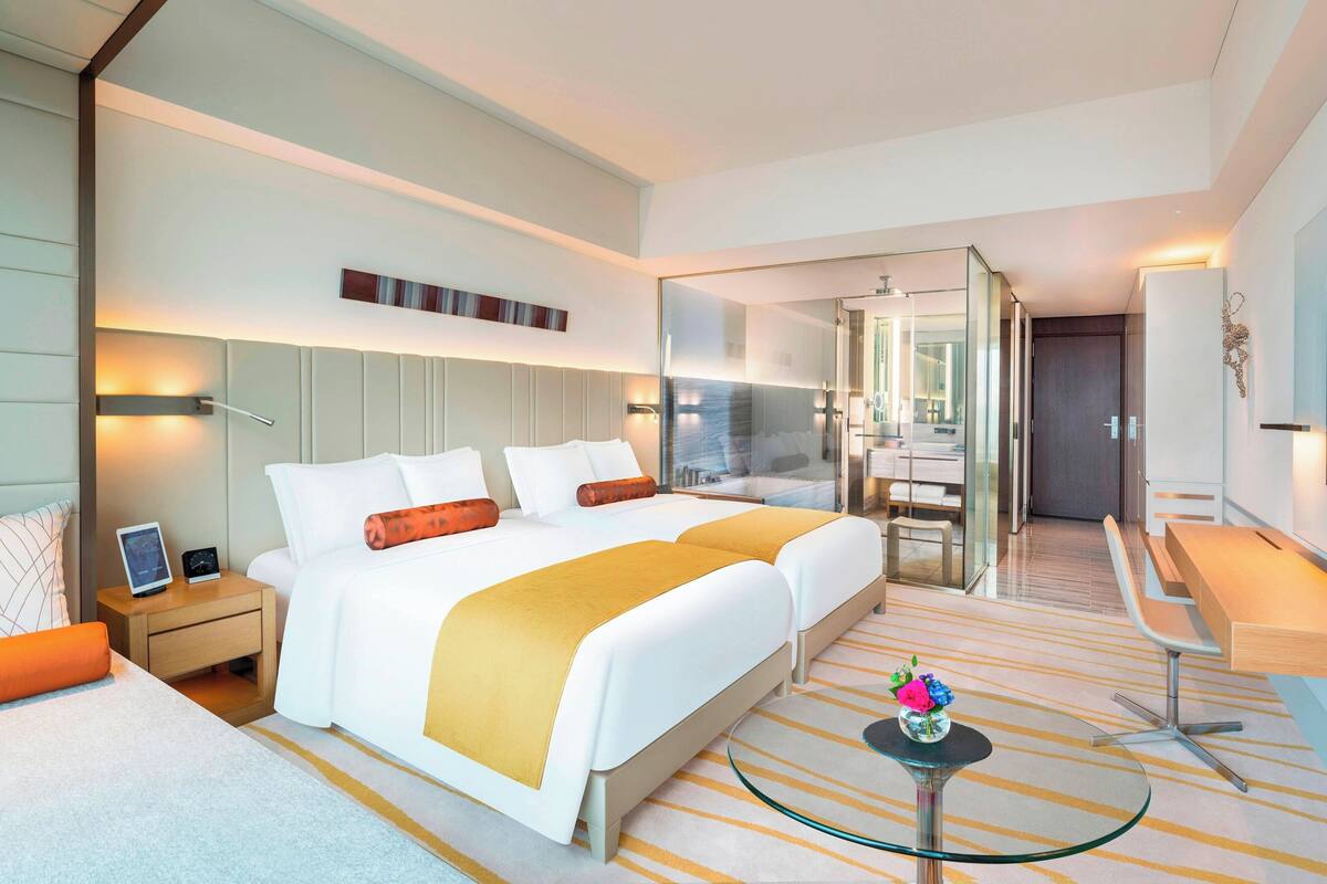A modern hotel room with a large bed, bedside tables, a glass coffee table, and a view of the bathroom.