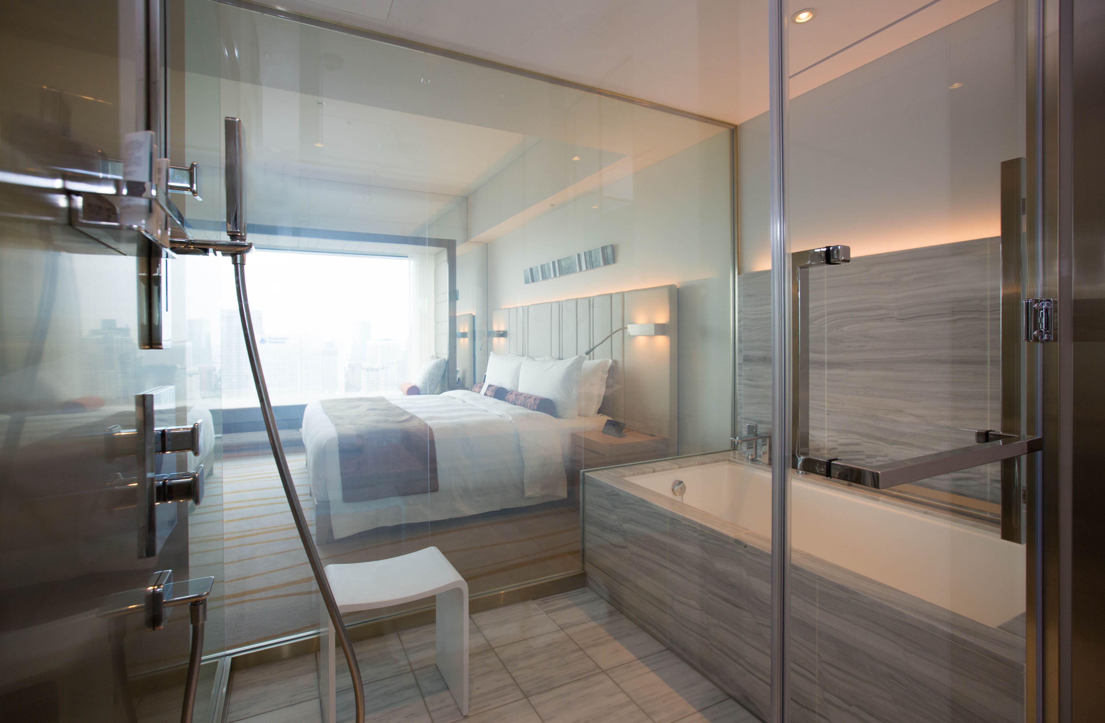 Una habitación de hotel moderna con cabina de ducha de vidrio, una cama grande con ropa blanca, una zona de estar con una mesita pequeña y un espejo grande sobre el lavabo.