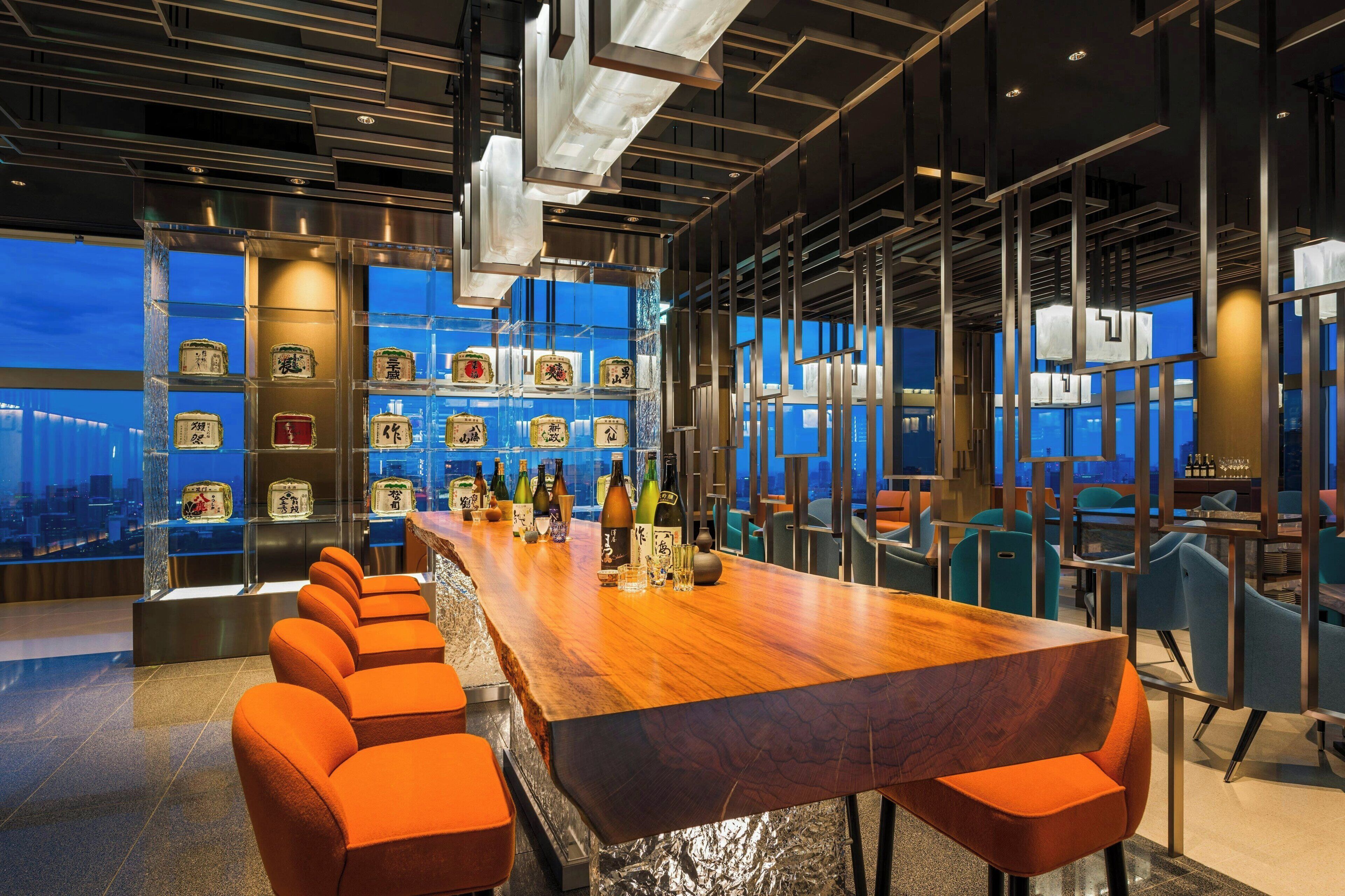 A modern bar area with a long wooden counter, orange seating, and a city view through large windows.