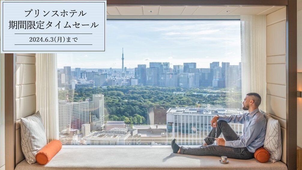 Una persona seduta su una panchina vicino a una grande finestra con vista su un paesaggio urbano con edifici moderni e un parco.