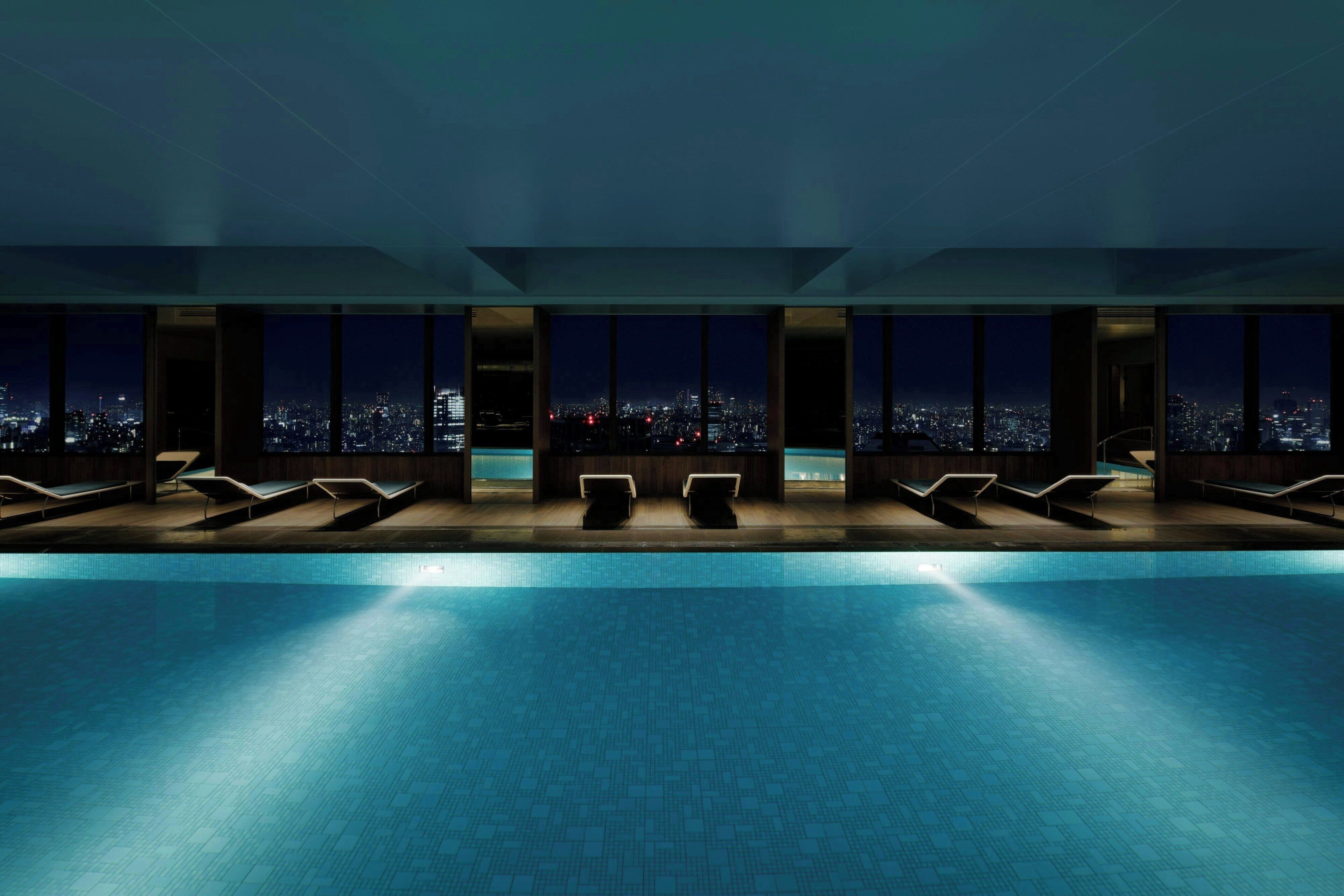 Piscina interior moderna con vista a la ciudad, varios sillones de sol y techo de vidrio.