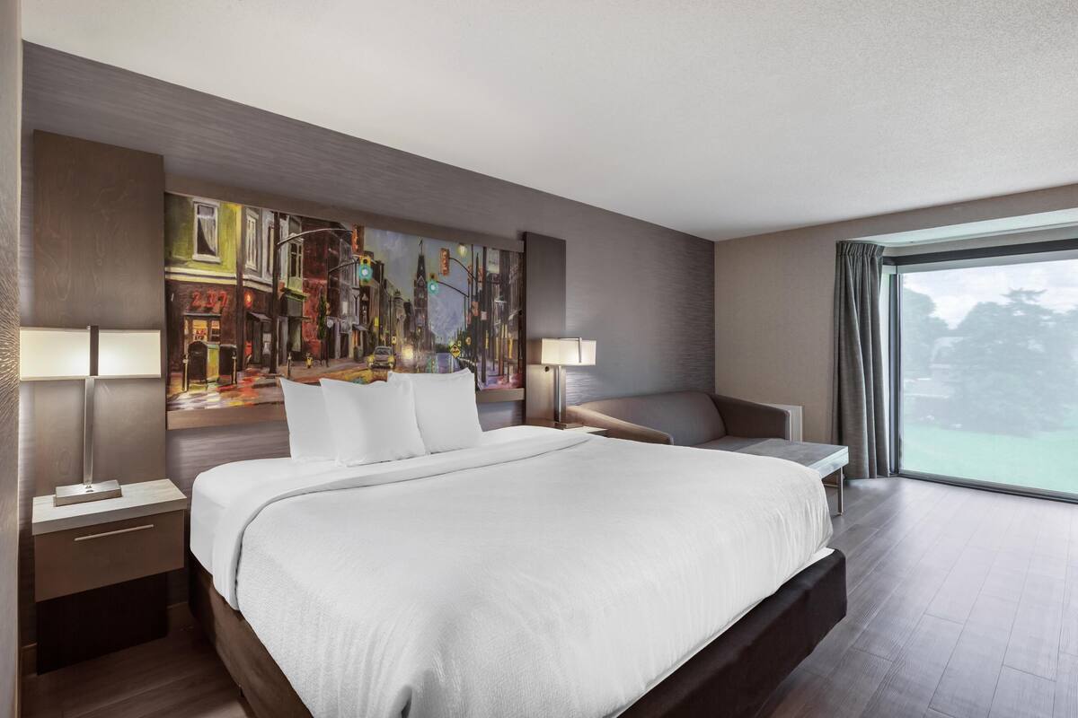 A modern hotel room with a large bed, bedside tables, a painting on the wall, and a view through the window.