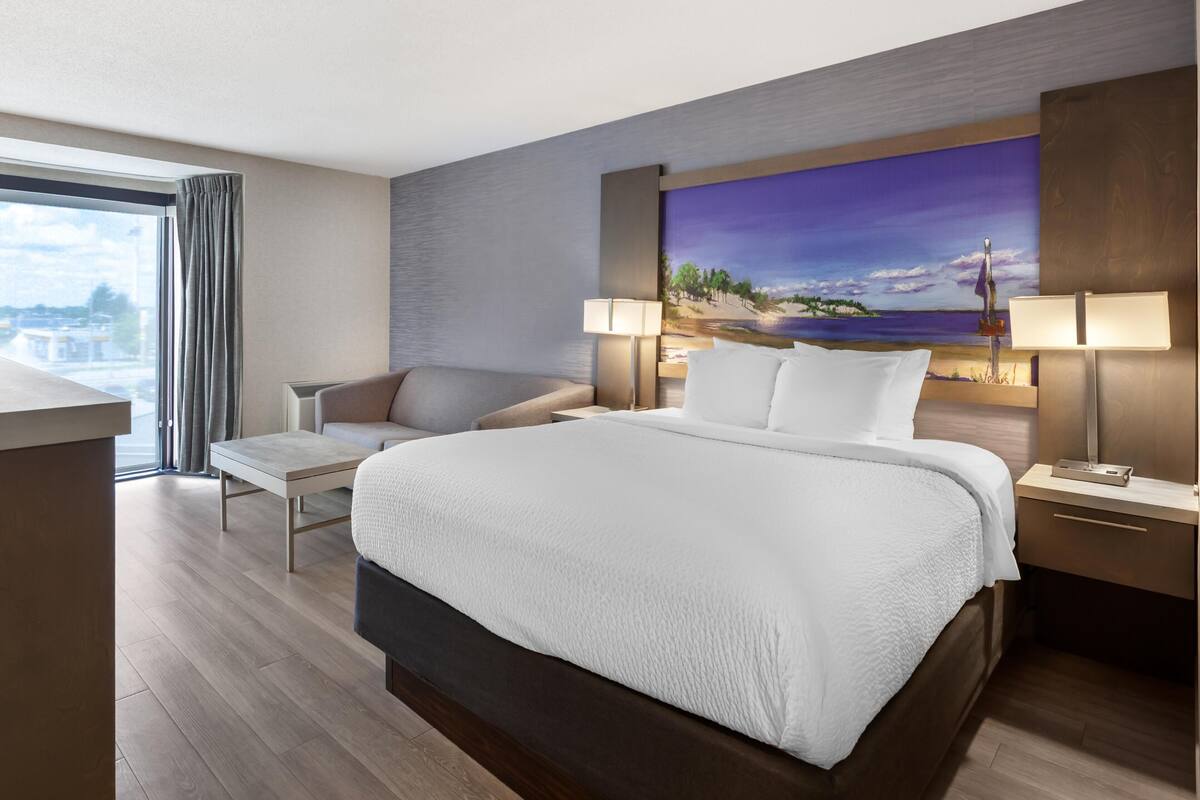 A modern hotel room with a large bed, a desk, a chair, and a painting on the wall.