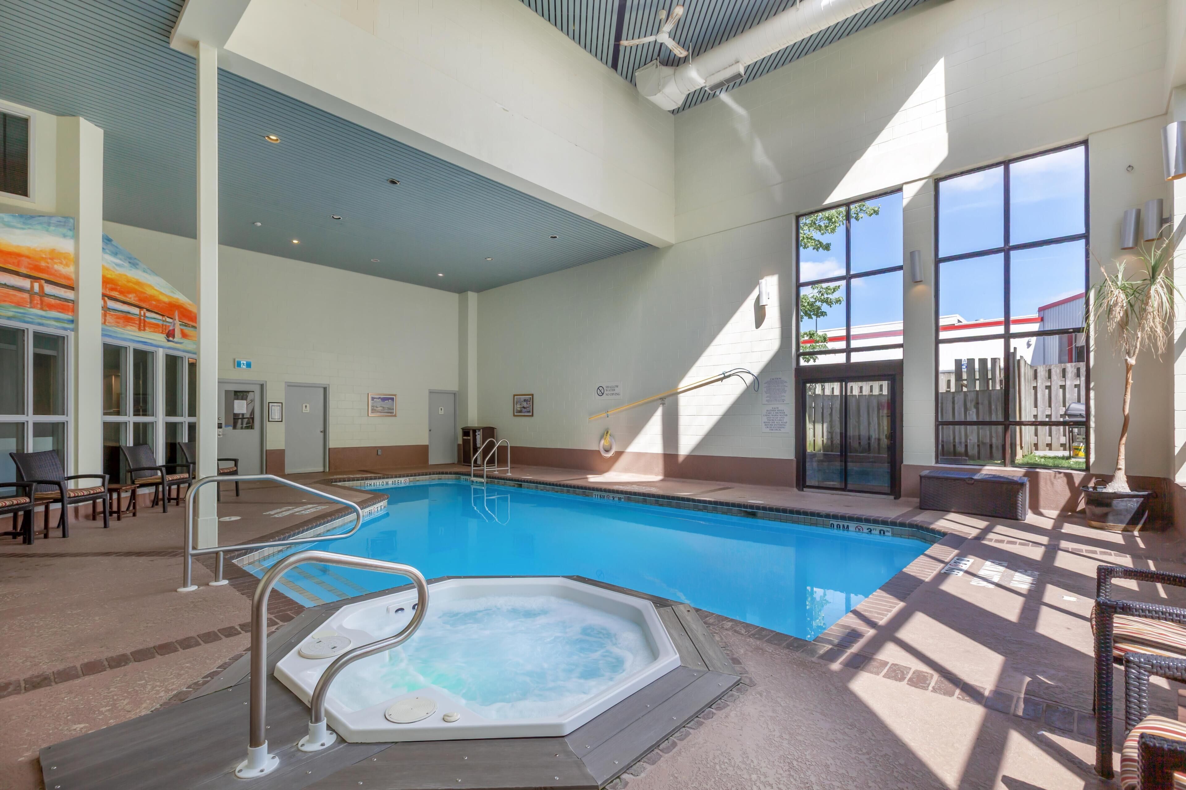 An indoor swimming pool with a hot tub, sunlit seating area, and large windows.