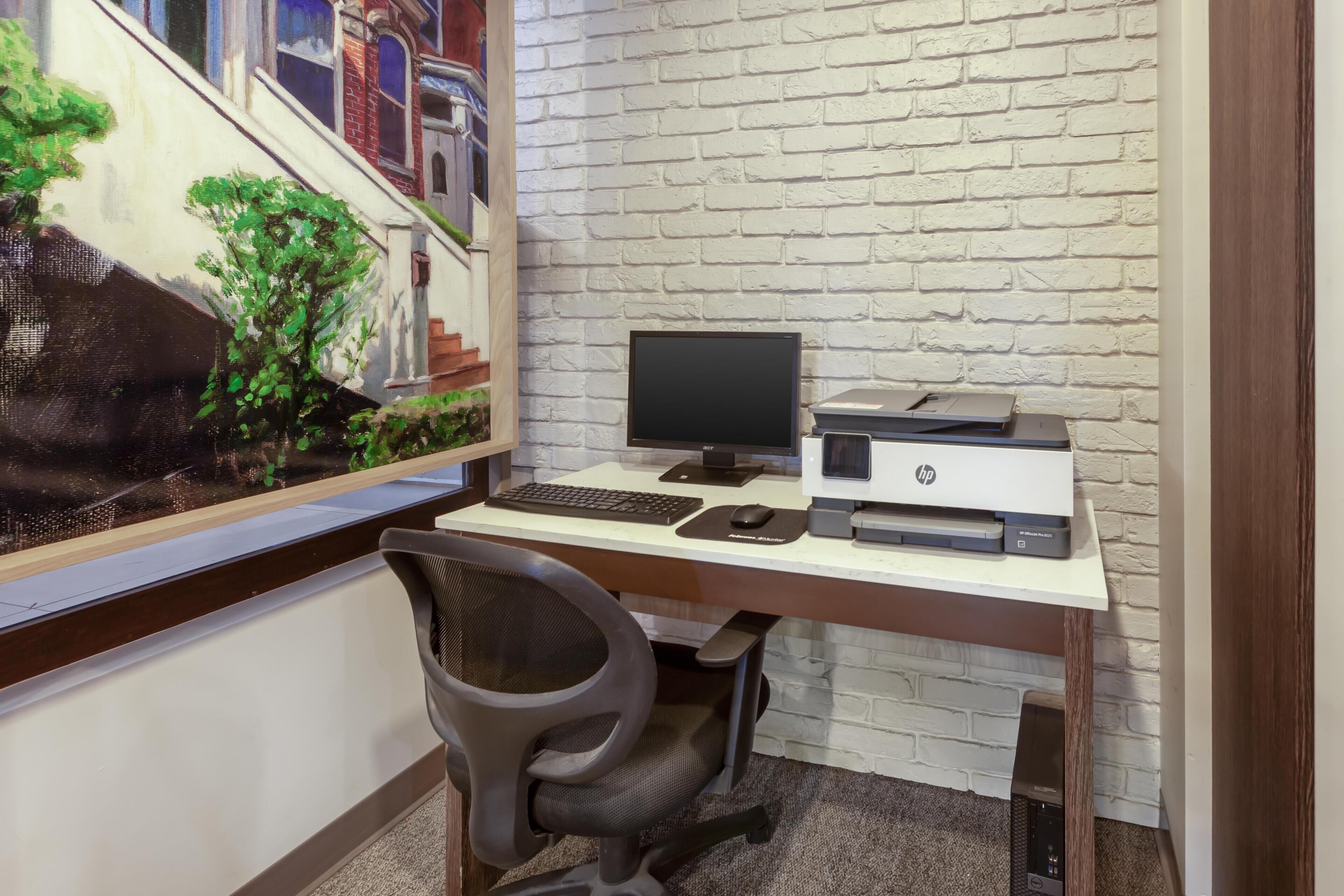A computer desk with a monitor, keyboard, mouse, and printer against a white brick wall.