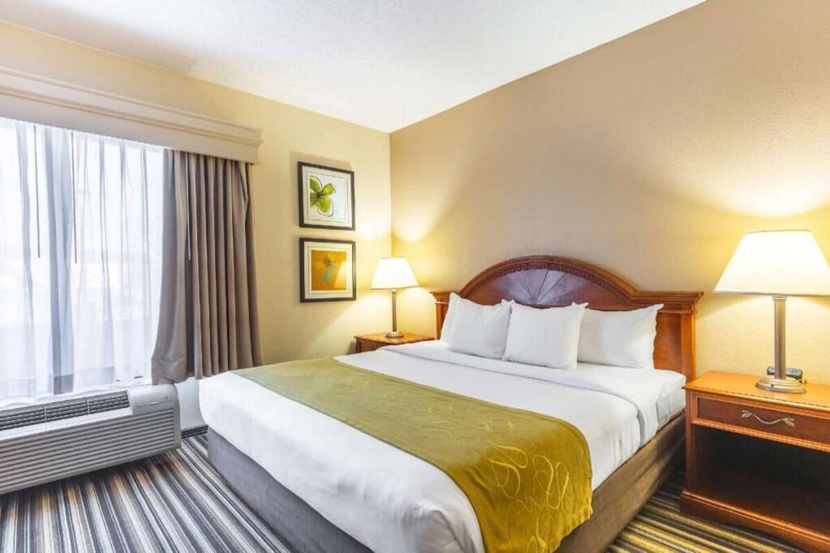 A hotel room with a bed, two bedside lamps, a window with curtains, a wall-mounted air conditioner, and two framed pictures.