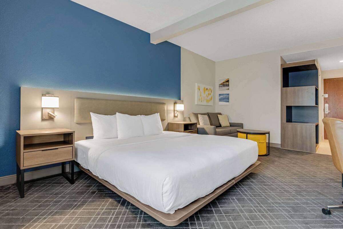 A modern hotel room with a large bed, bedside tables with lamps, a sofa, and a built-in shelving unit.