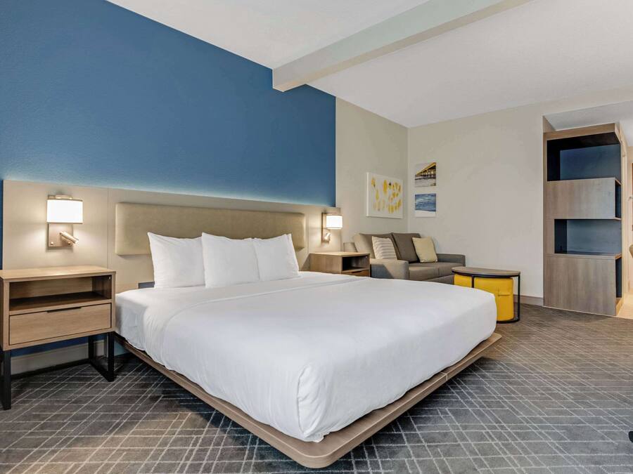 A modern hotel room with a large bed, bedside tables with lamps, a sofa, and a built-in shelving unit.