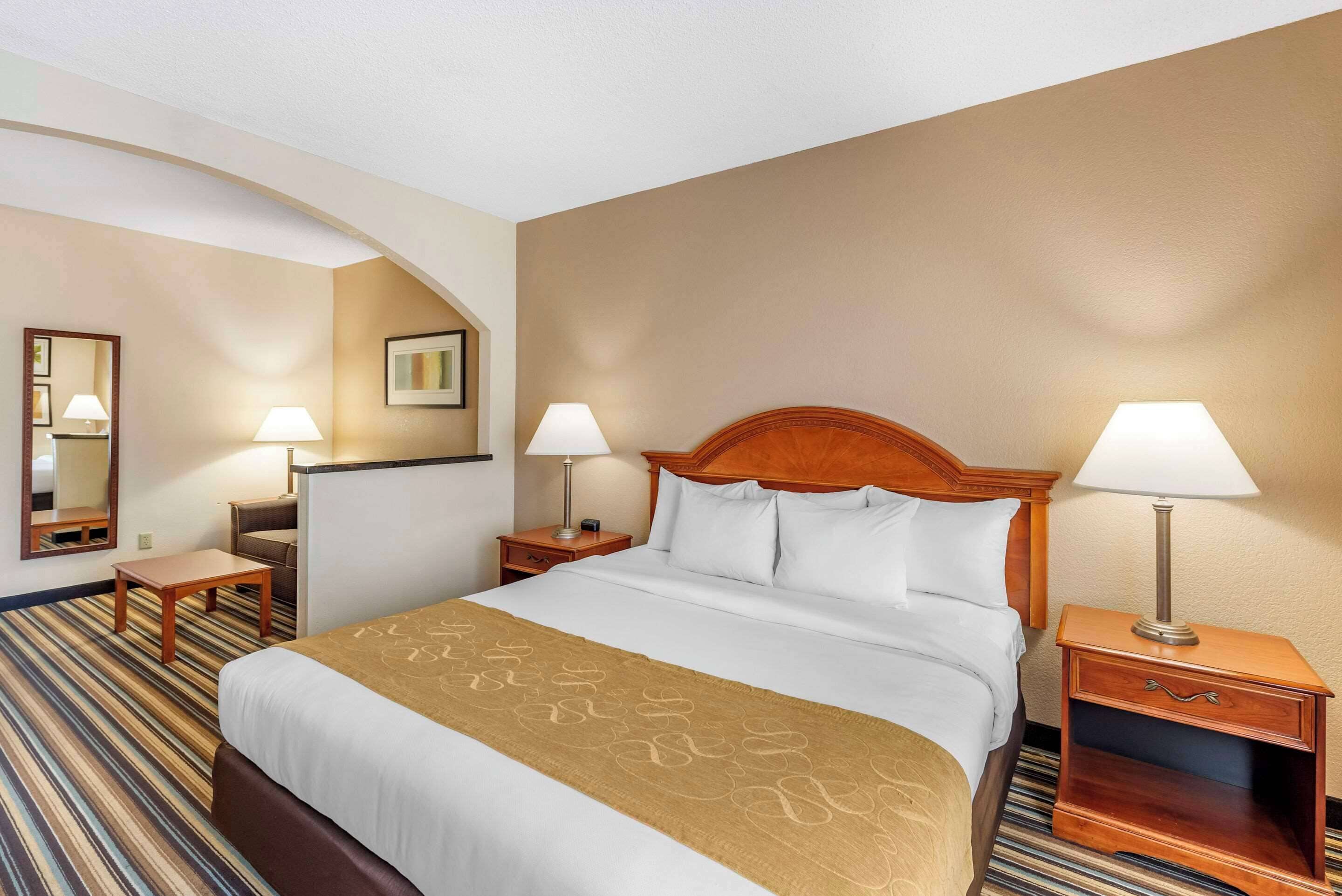 A hotel room with a large bed, two bedside tables with lamps, a wooden headboard, a striped carpet, and a wall-mounted mirror.