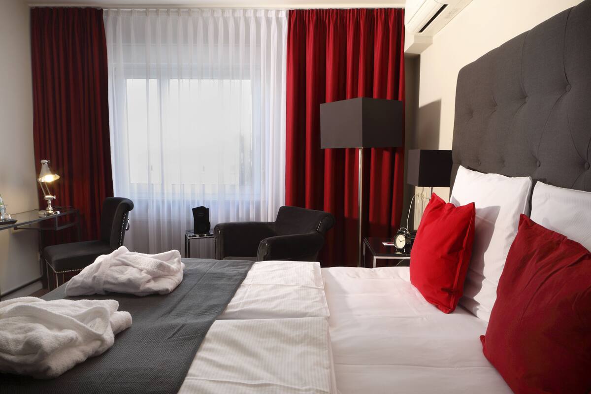 Habitación de hotel con cama, dos sillones, escritorio con lámpara y ventana grande con cortinas rojas.