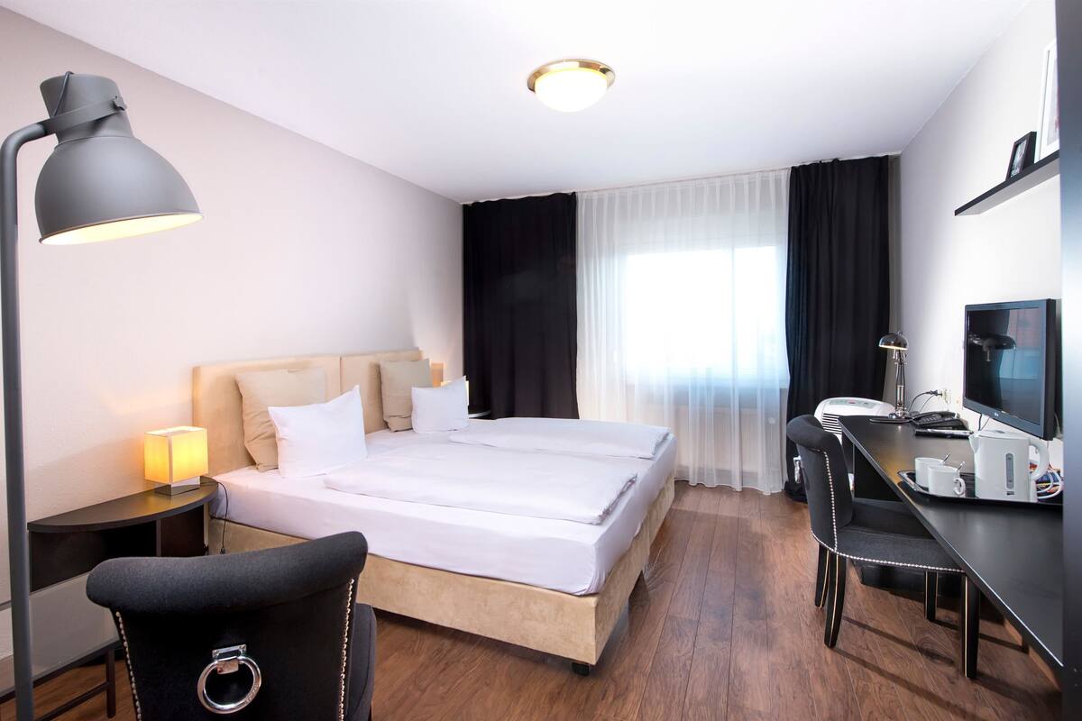 Habitación de hotel con cama, escritorio, silla, lámpara de escritorio, televisión y mesita de noche con lámpara.