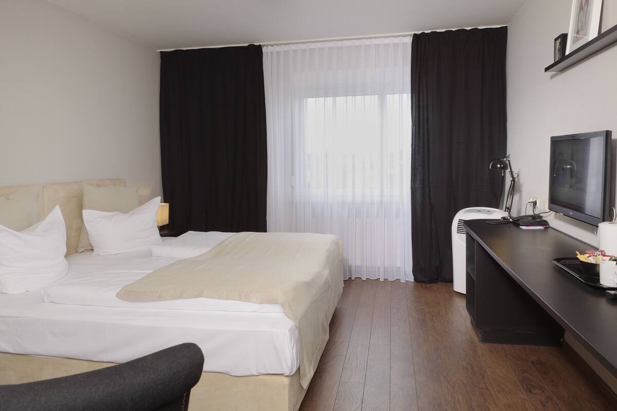 Una habitación de hotel con una cama, un escritorio con televisor y una ventana con cortinas translúcidas.