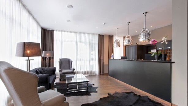 A modern living room with a sofa, armchair, coffee table, and a black leather rug. There is a desk with a computer and a lamp. - TRYP by Wyndham Bremen Airport (Bremen)