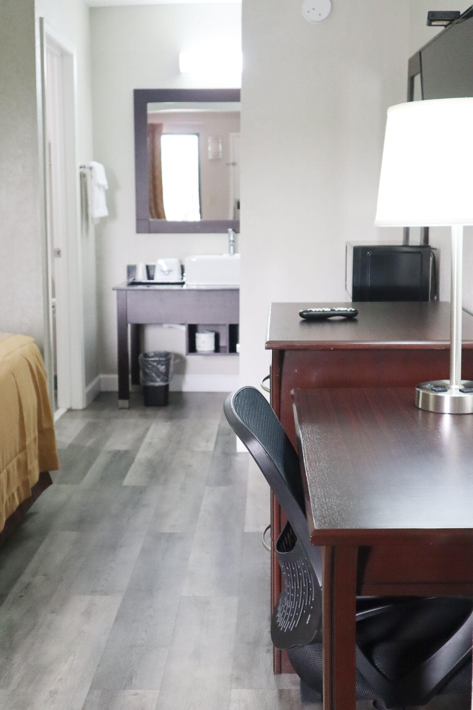 A hotel room with a bed, a desk with a lamp, a chair, a TV, and a bathroom visible in the background.