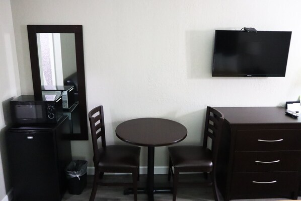 A compact hotel room with a single round table, two chairs, a small black refrigerator, a microwave, a TV mounted on the wall, and a dark wooden dresser with drawers.