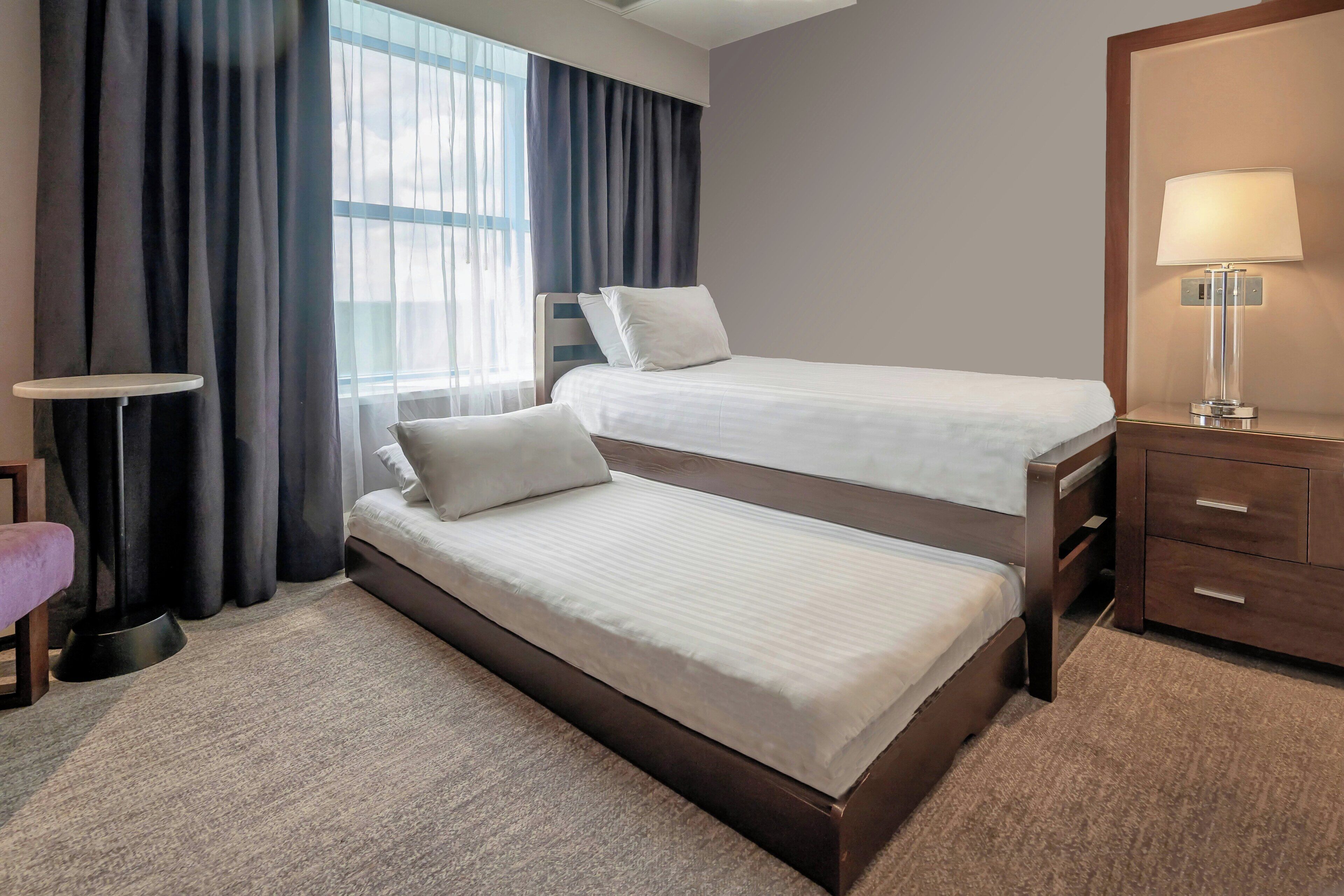 Habitación de hotel con dos camas, una mesita de noche, una lámpara, una silla y un ventanal con cortinas.