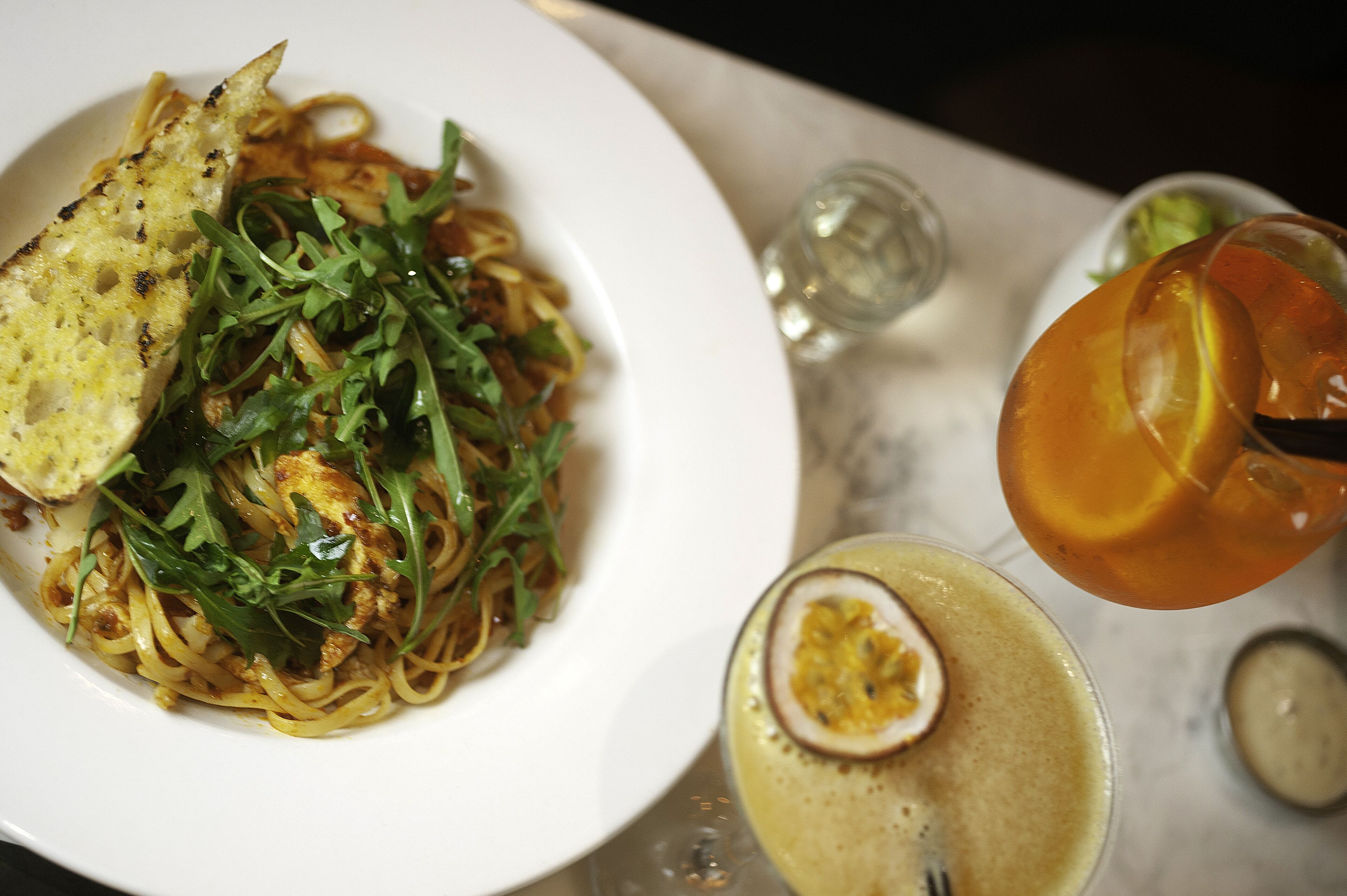 Un plato de pasta con verduras, una rebanada de pan y un vaso de bebida de color ámbar, junto con un recipiente pequeño de salsa.
