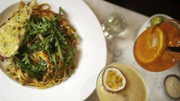 A plate of pasta with greens and a slice of bread, a glass of amber-colored drink, and a small dish of sauce.