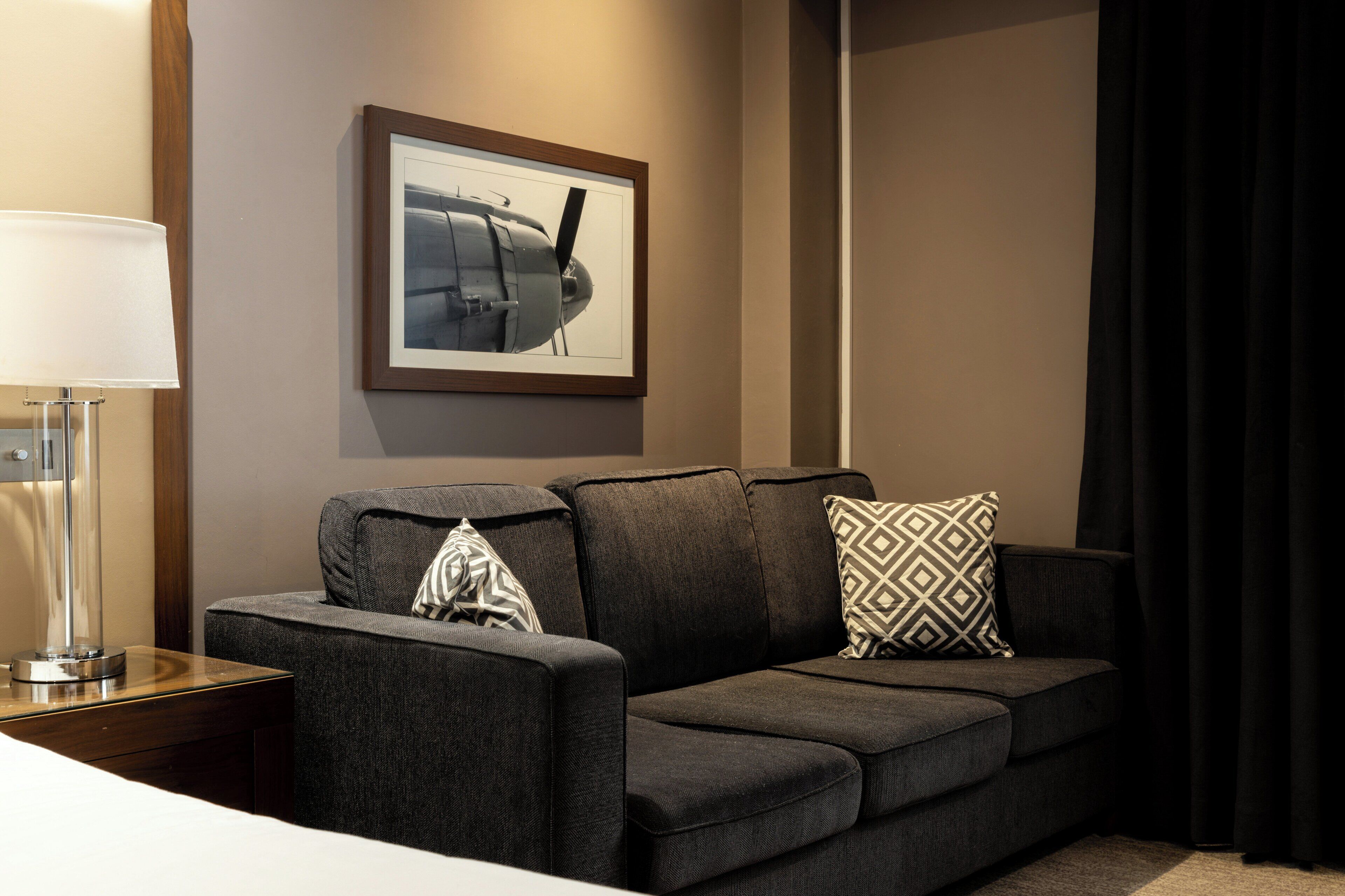 A modern hotel room with a dark gray sofa, a framed picture on the wall, a bedside lamp, and a dark curtain.