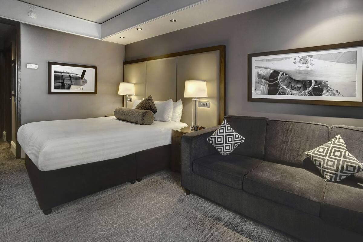 A hotel room with a large bed, a sofa, two wall-mounted lamps, and two framed pictures.