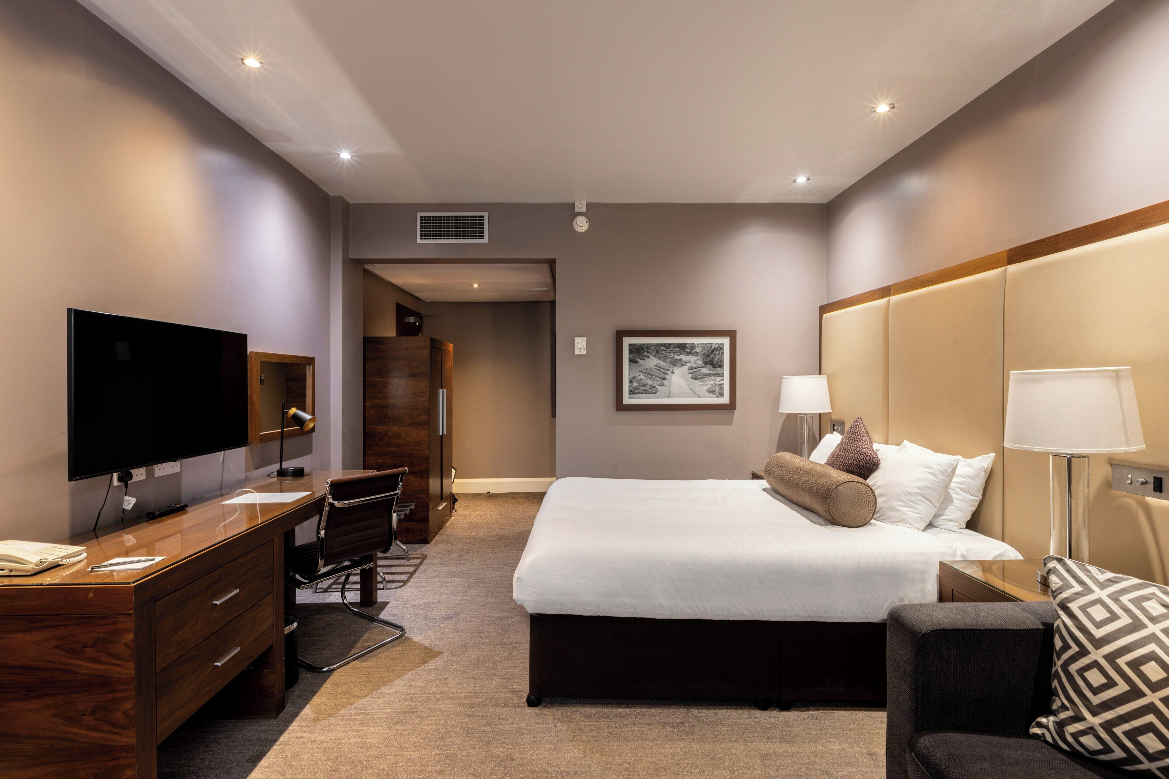A hotel room with a bed, desk, TV, and a framed picture on the wall.