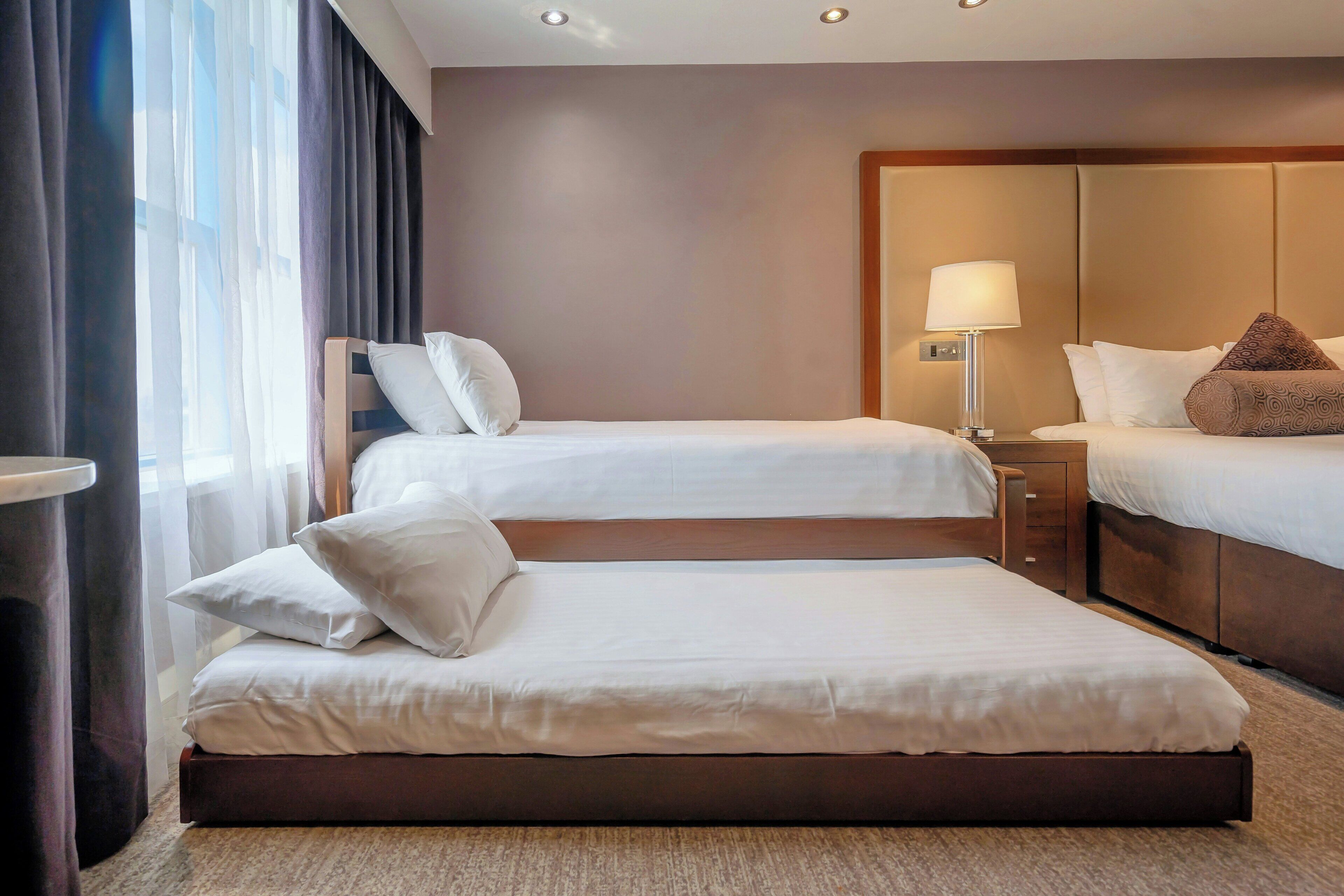 Habitación de hotel con dos camas, una mesita de noche con lámpara y un ventanal con cortinas.