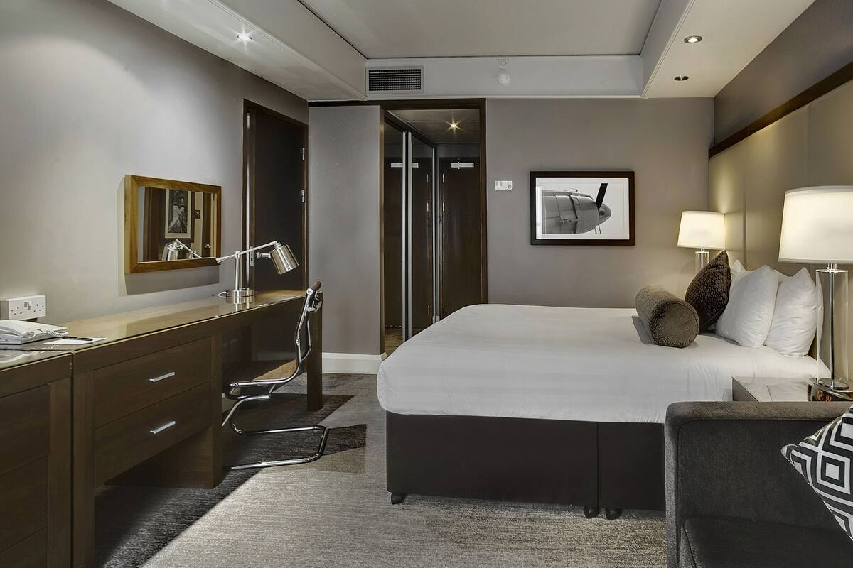 A hotel room with a bed, bedside table, lamp, desk, chair, and a mirror.