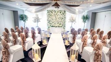 Salón de bodas con filas de sillas blancas, un pasillo blanco y decoraciones florales en la pared.