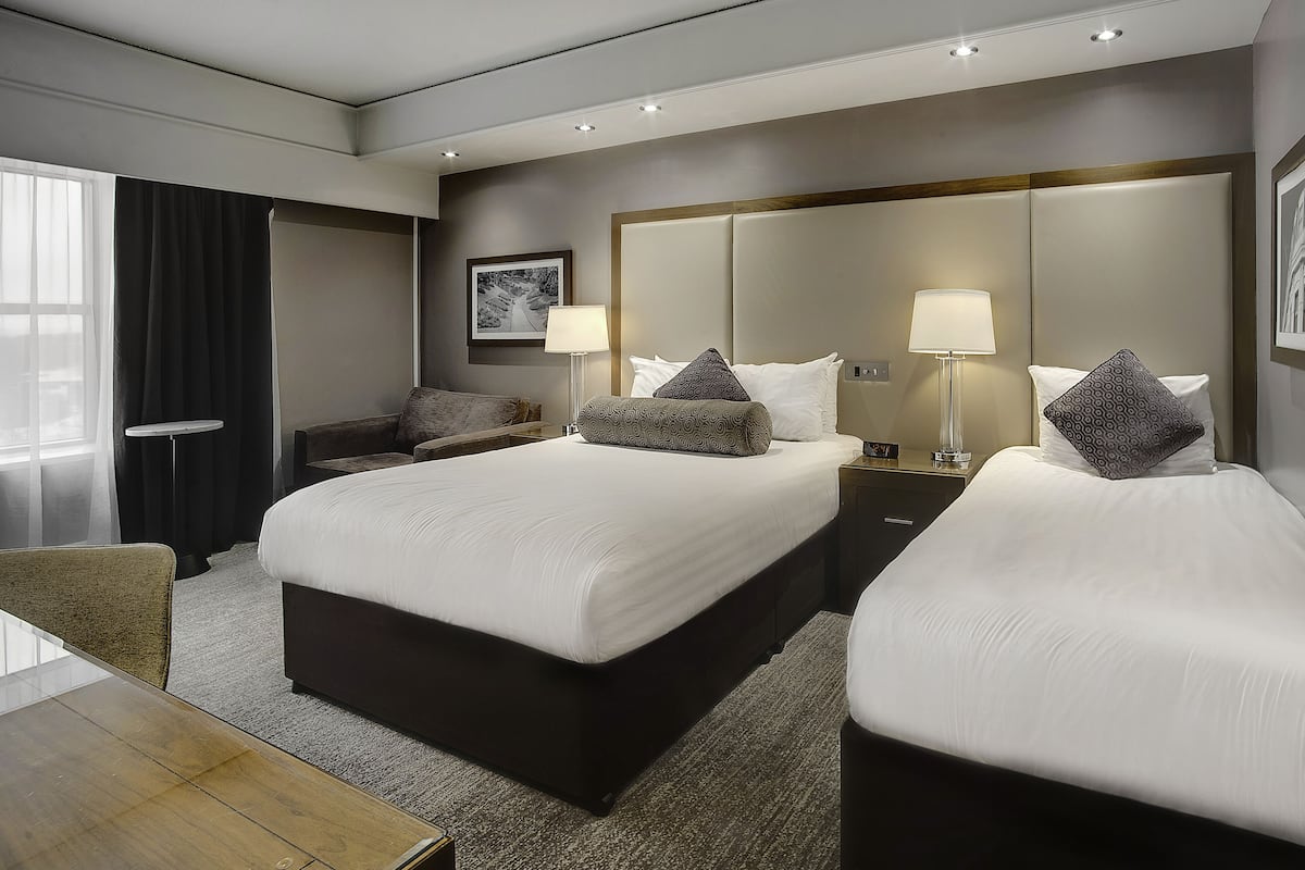 A hotel room with two beds, a small table, a chair, and a bedside table with lamps.