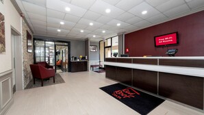 A reception area with a flat-screen TV displaying 'Free Wi-Fi All Rooms,' a reception desk, a seating area with chairs, and a doormat with 'Root Room' text. - Red Roof Inn Petersburg - Fort Lee (Petersburg)