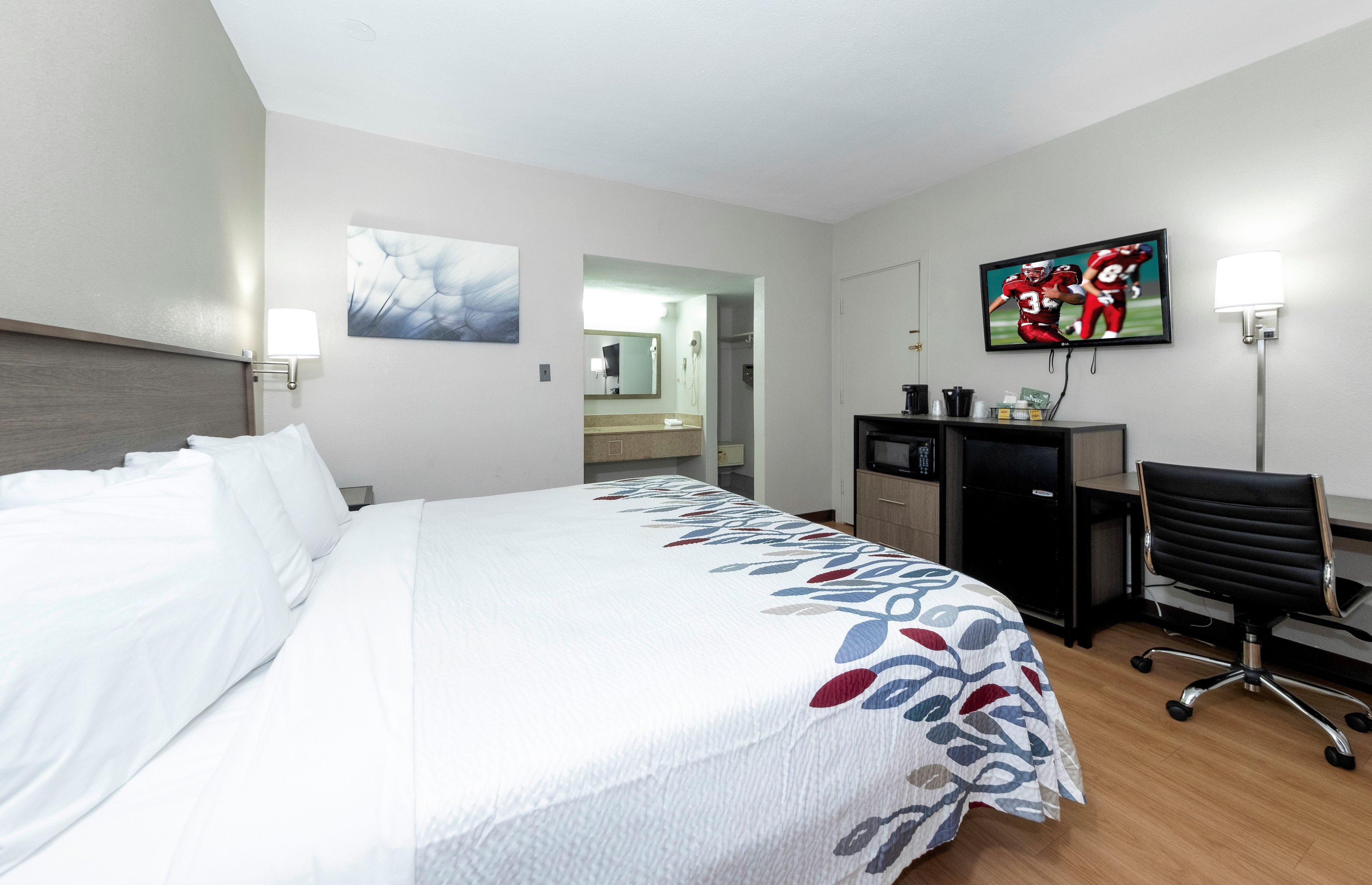 A hotel room with a bed, a desk, a chair, a television, and a bathroom visible through an open door.