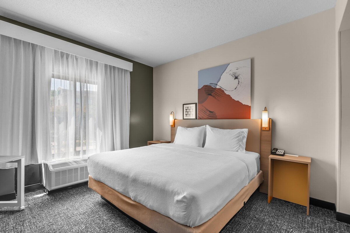 A hotel room with a large bed, a window with sheer curtains, a wall-mounted artwork, and bedside lamps.