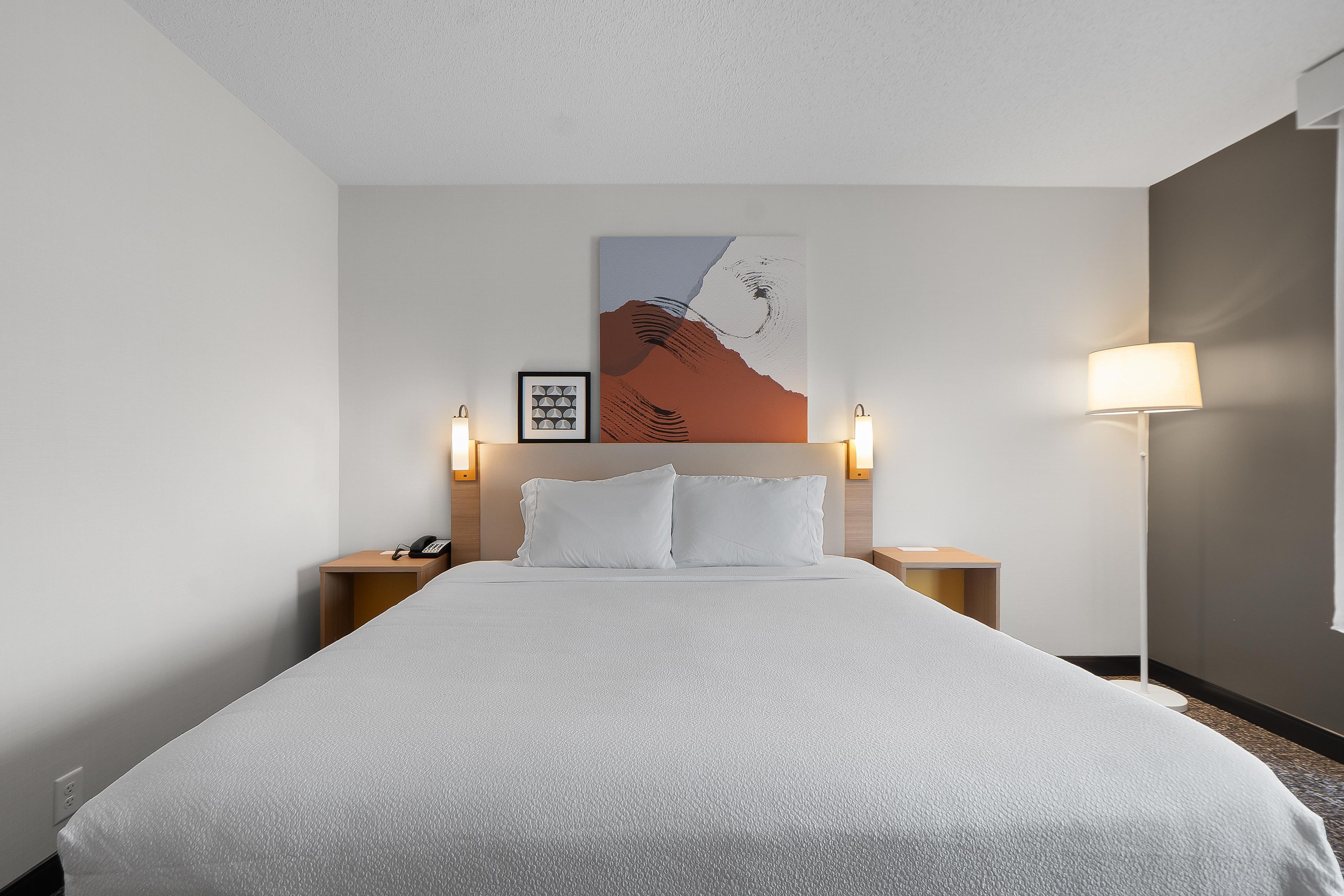 A hotel room with a large bed, two bedside lamps, a framed artwork on the wall, and a telephone on the nightstand.