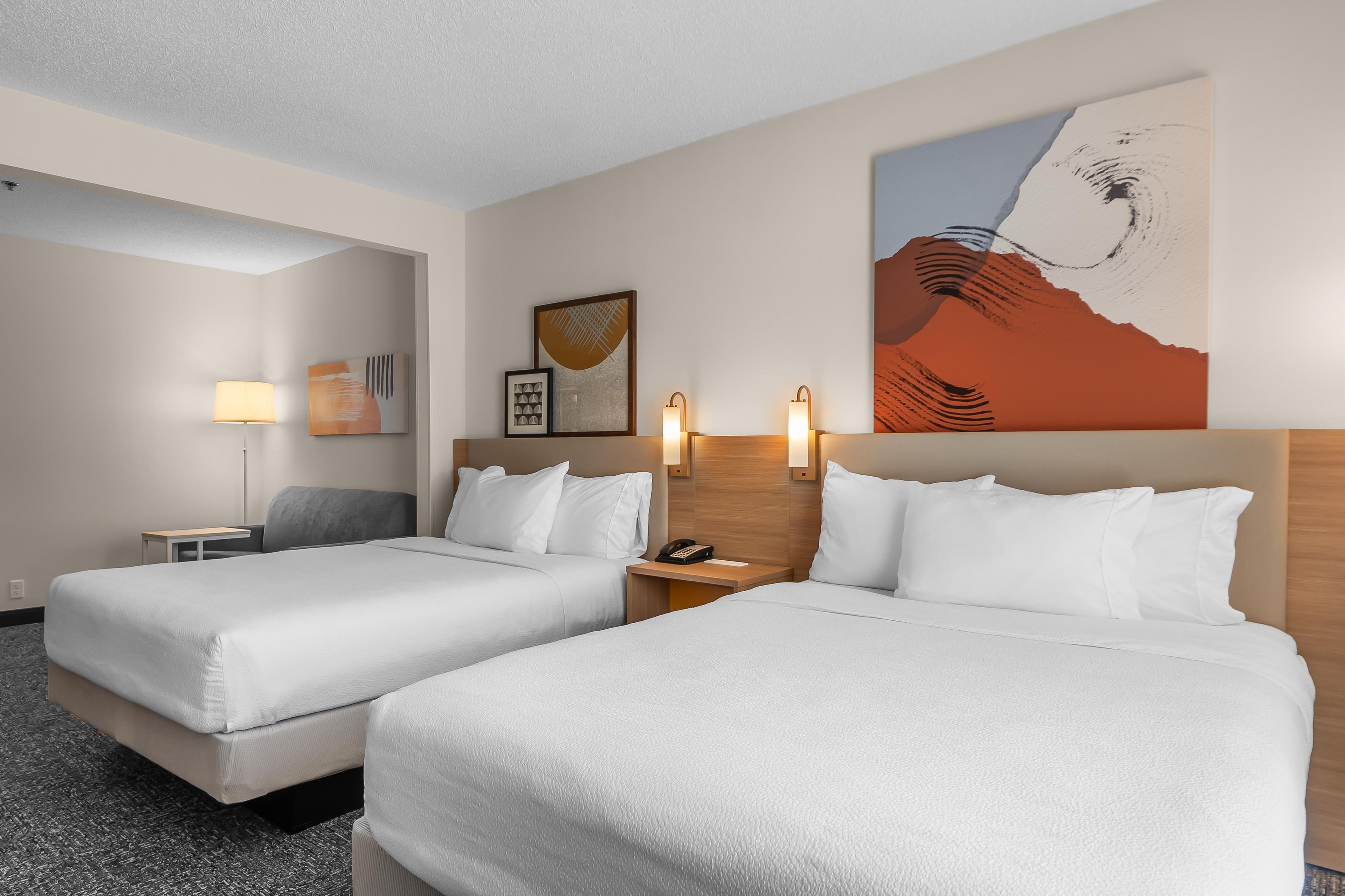 A hotel room with two beds, a nightstand, a lamp, and artwork on the walls.
