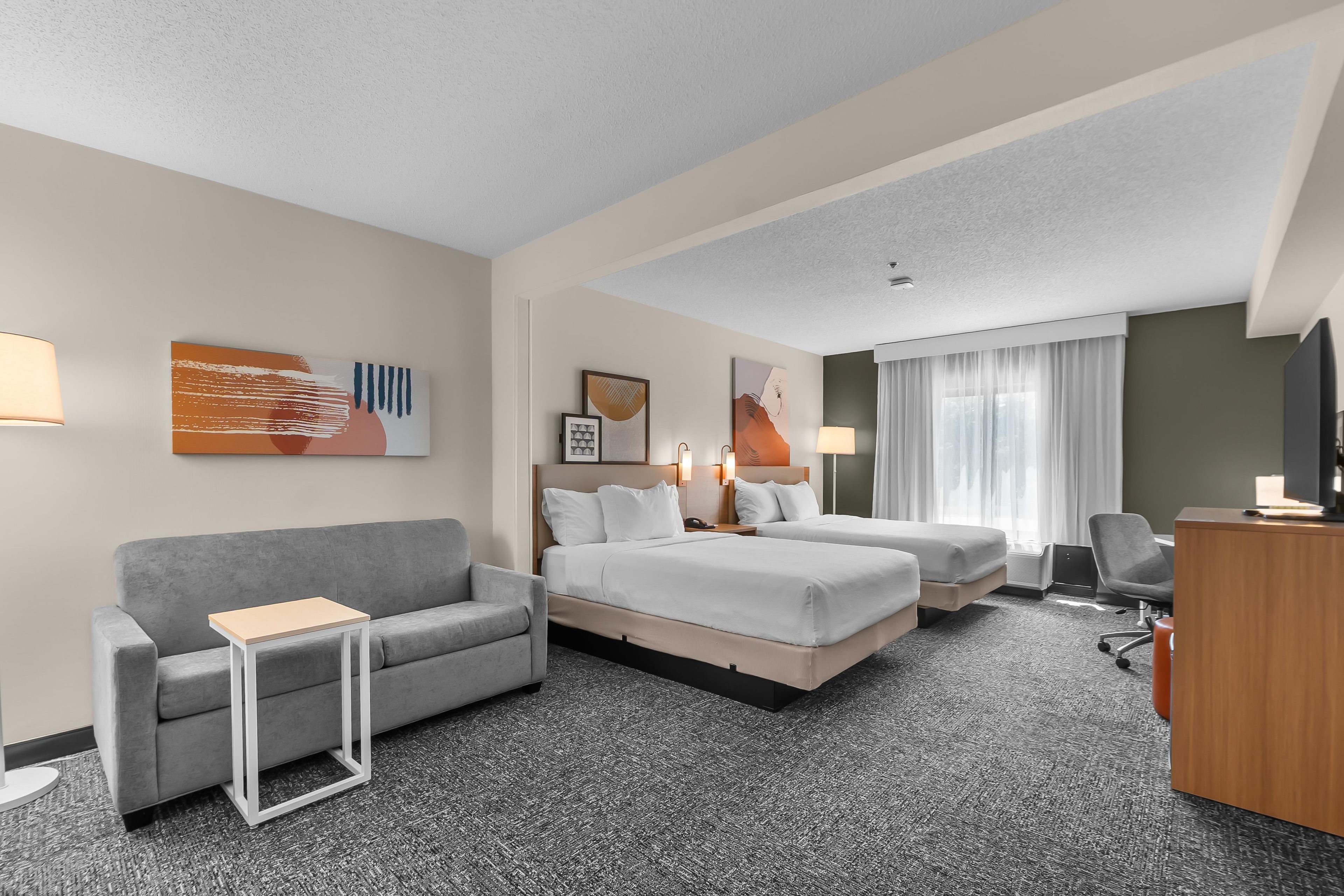 A hotel room with a grey sofa, two single beds, a desk with a chair, and a television.