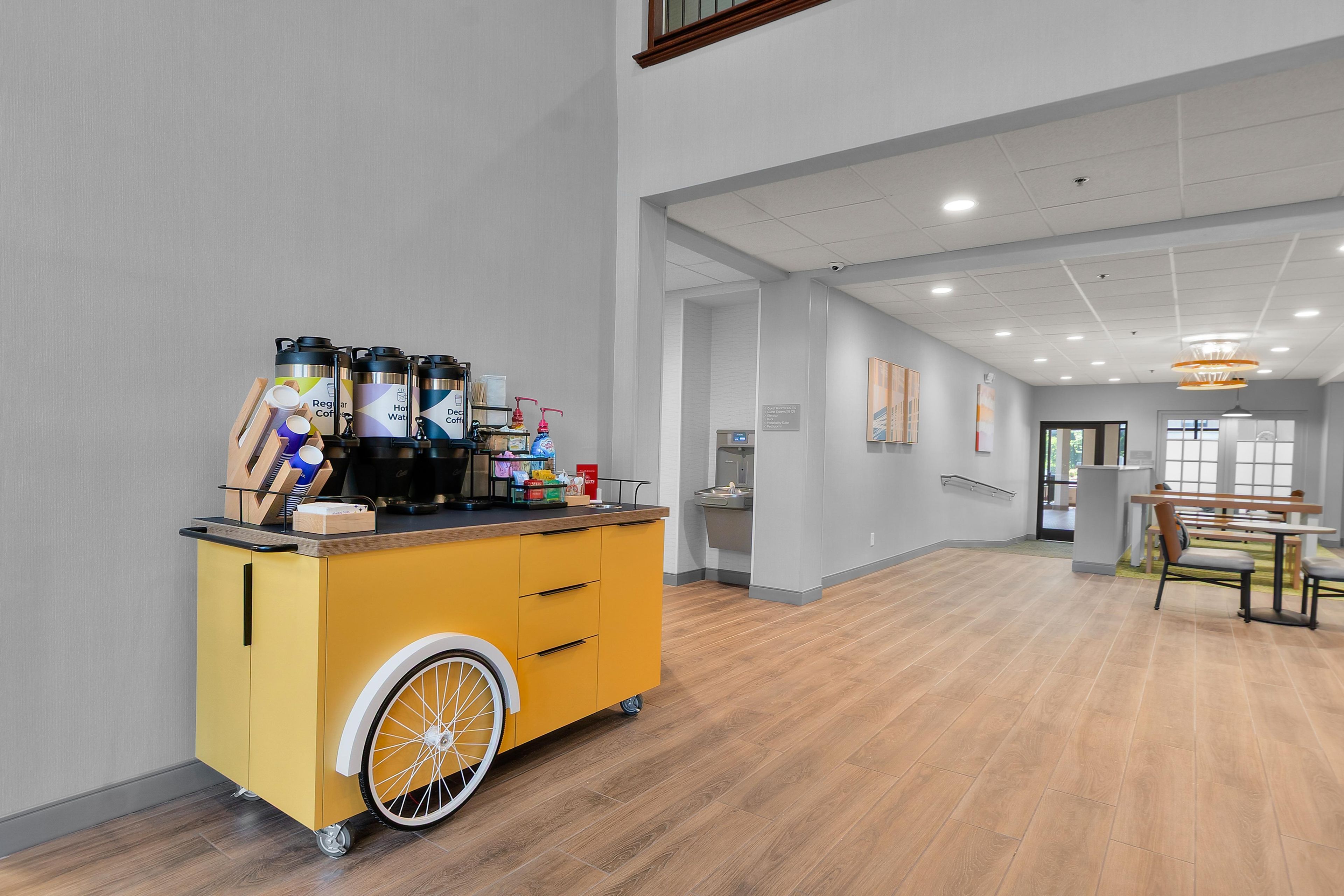 A hotel room with a yellow cart containing coffee and tea dispensers, a sink, and a bench.