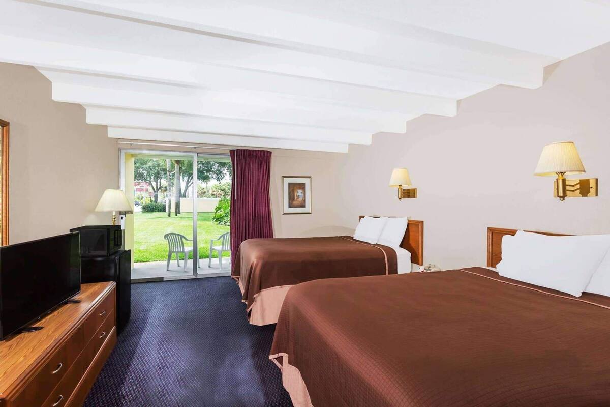 A hotel room with two beds, a TV, a desk, and a sliding glass door to a garden.
