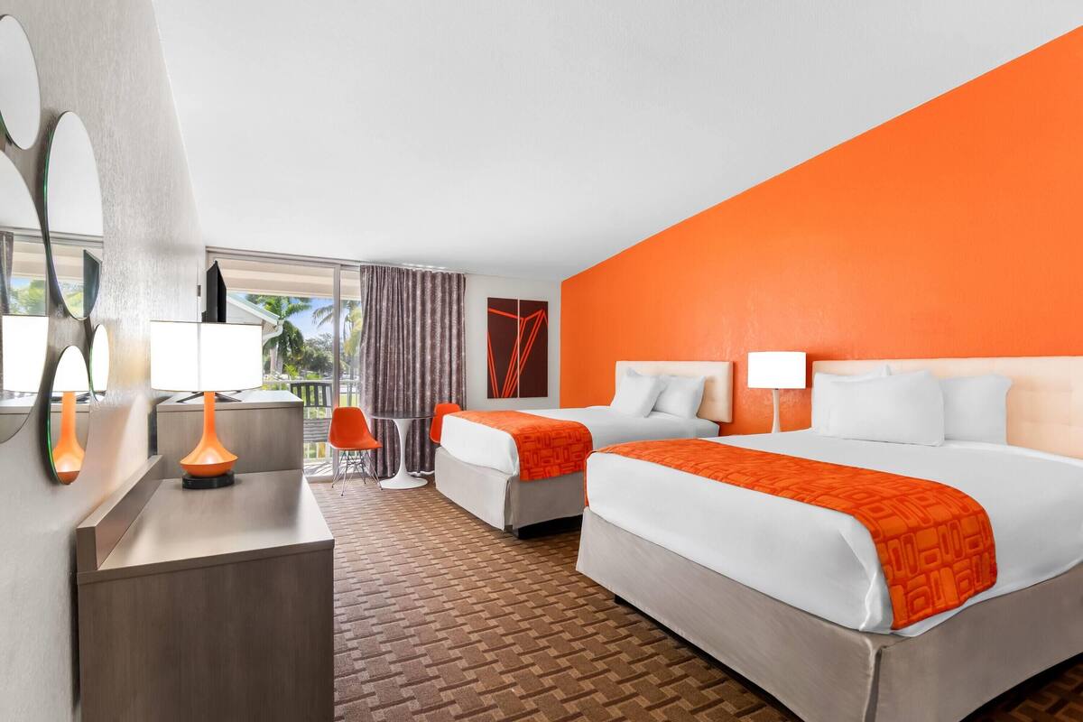 A hotel room with two beds, a desk with a lamp, a chair, and a view of palm trees.
