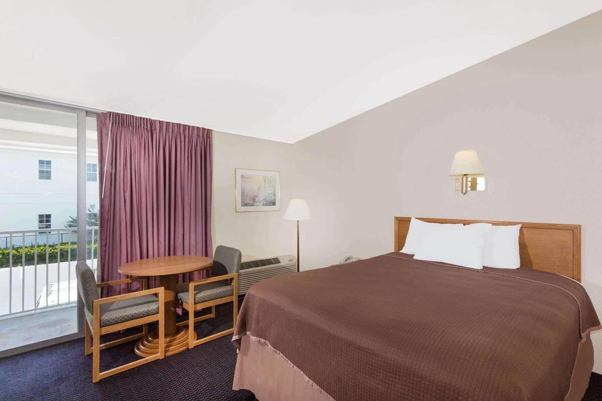 A hotel room with a bed, a desk with two chairs, a window with curtains, a wall-mounted lamp, and a painting.