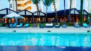 Zona de piscina con sillones de descanso y sombrillas, rodeada de edificios con balcones y terrazas.