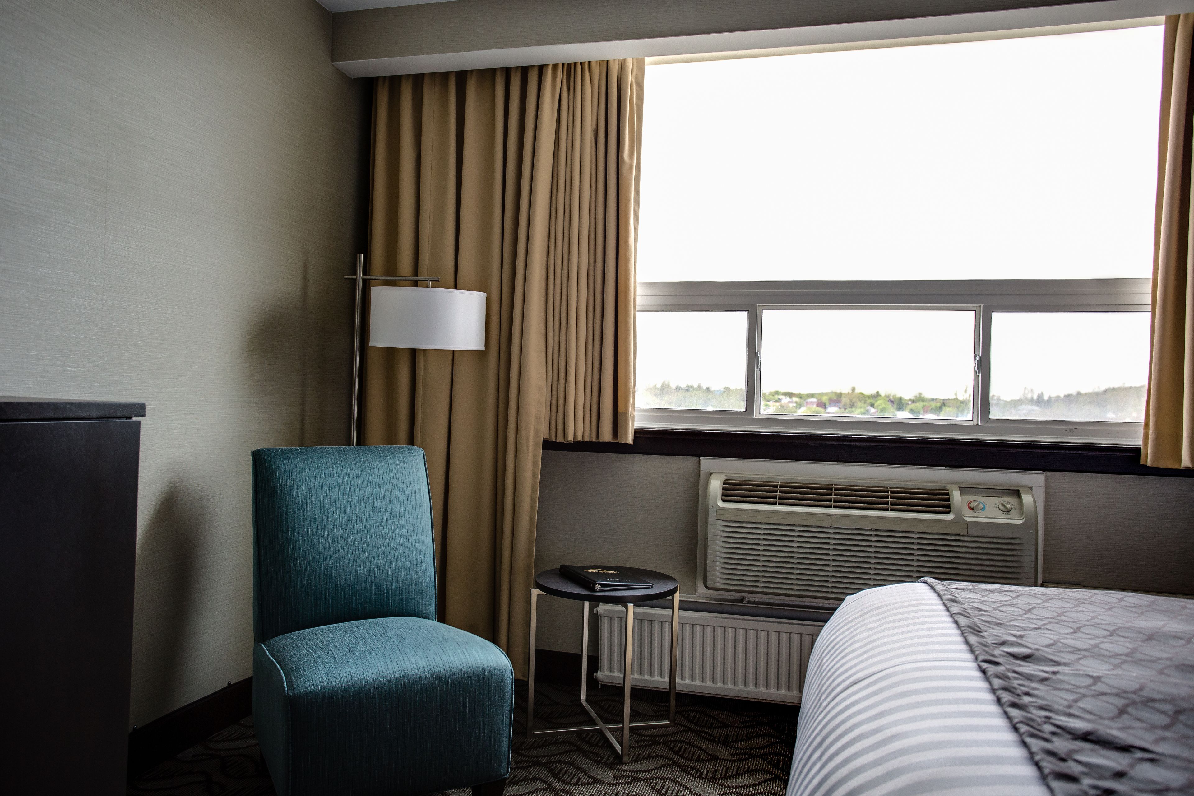 Ett hotellrum med en säng, en stol, ett bord, en lampa och ett fönster med gardiner.