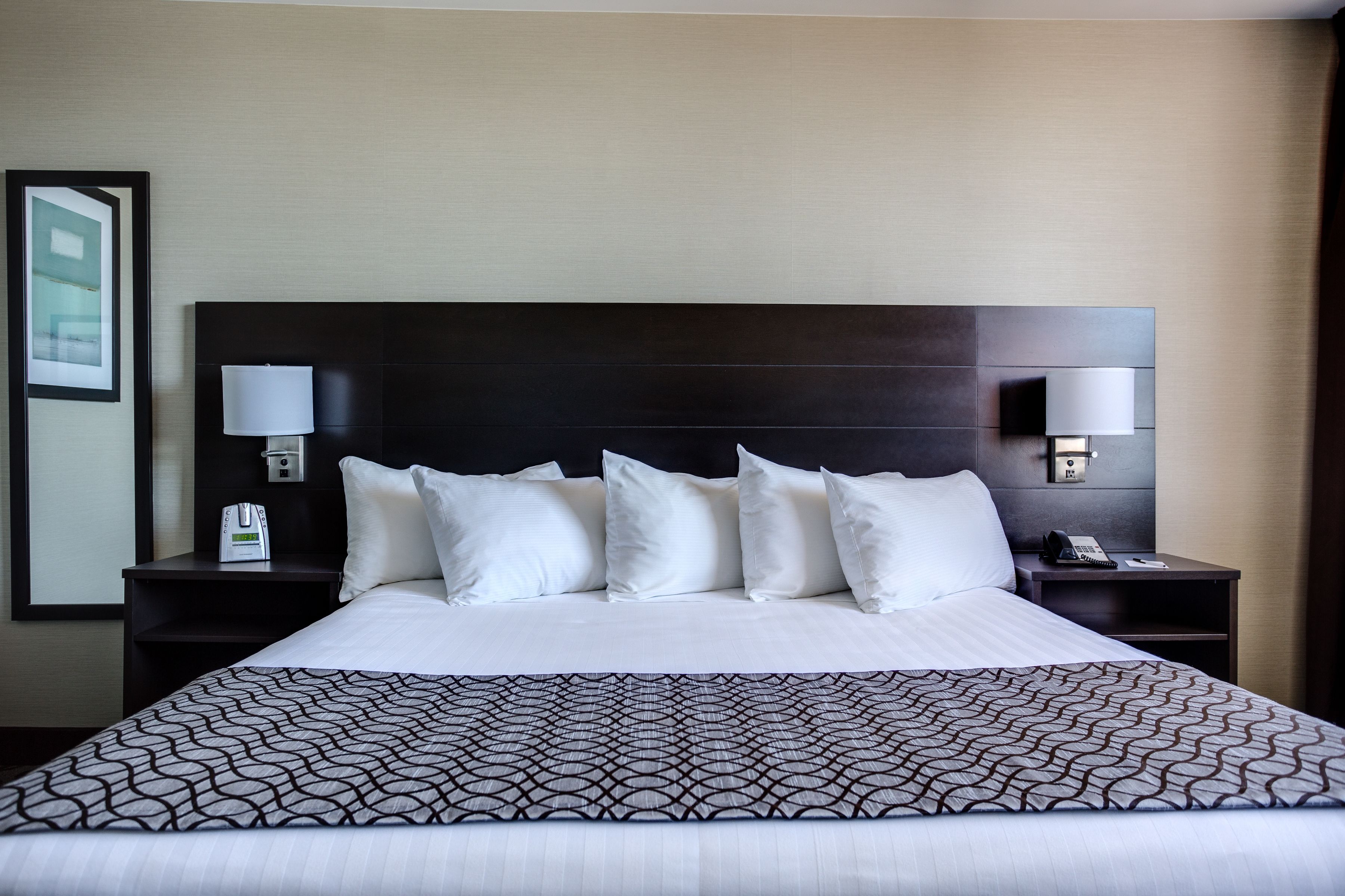 A hotel room with a large bed, two bedside tables with lamps, a framed picture on the left wall, and a dark headboard.